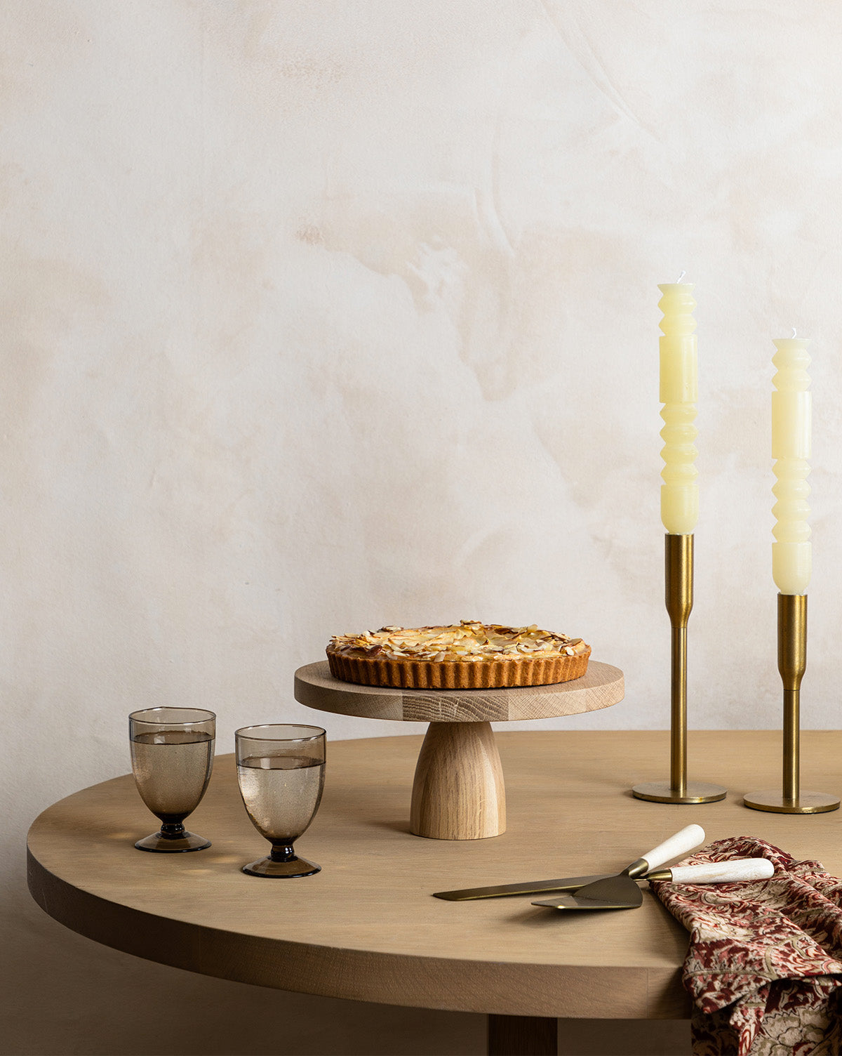 A Wooden Cake Stand holds a round tart, enhancing the table set with two smoky glasses, gold and white utensils, a patterned cloth, and two ivory spiral candles in gold holders, by McGee & Co.