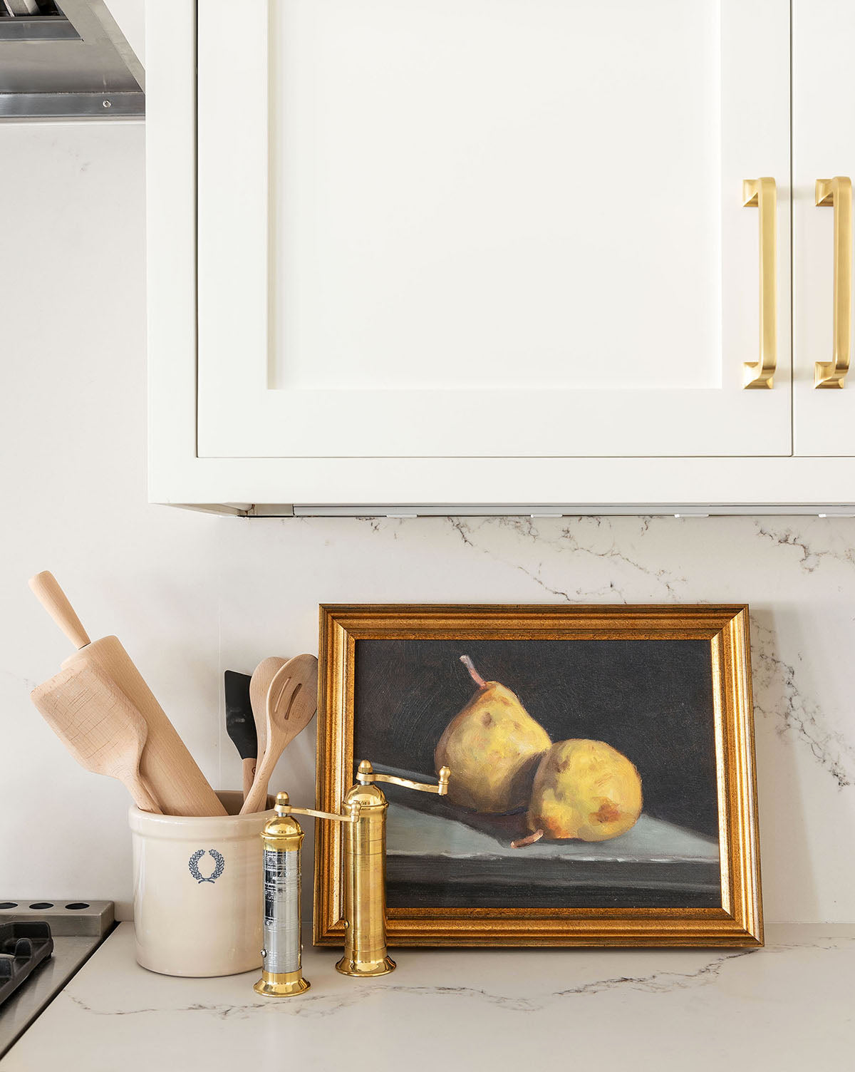 A kitchen counter features a gold salt grinder, ceramic utensil holder, and the framed Still Life with Pears artwork beneath white cabinets with gold handles, creating a charming, gallery wall-inspired space.
