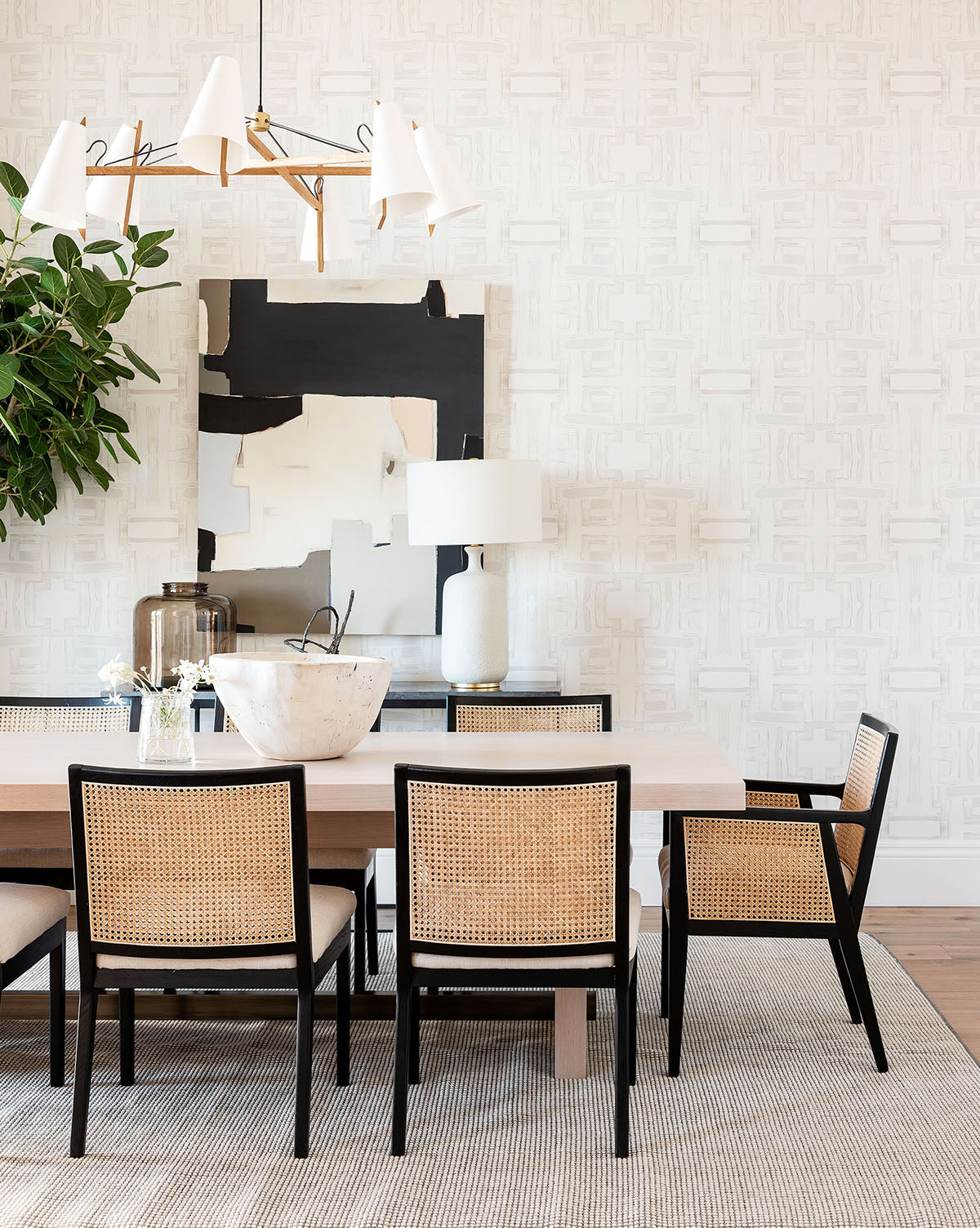 A modern dining room with a light wood table, six Landon Dining Chairs with cane backs and high-performance fabric, abstract wall art, a white lamp, potted plant, white chandelier, and a neutral rug by McGee & Co.