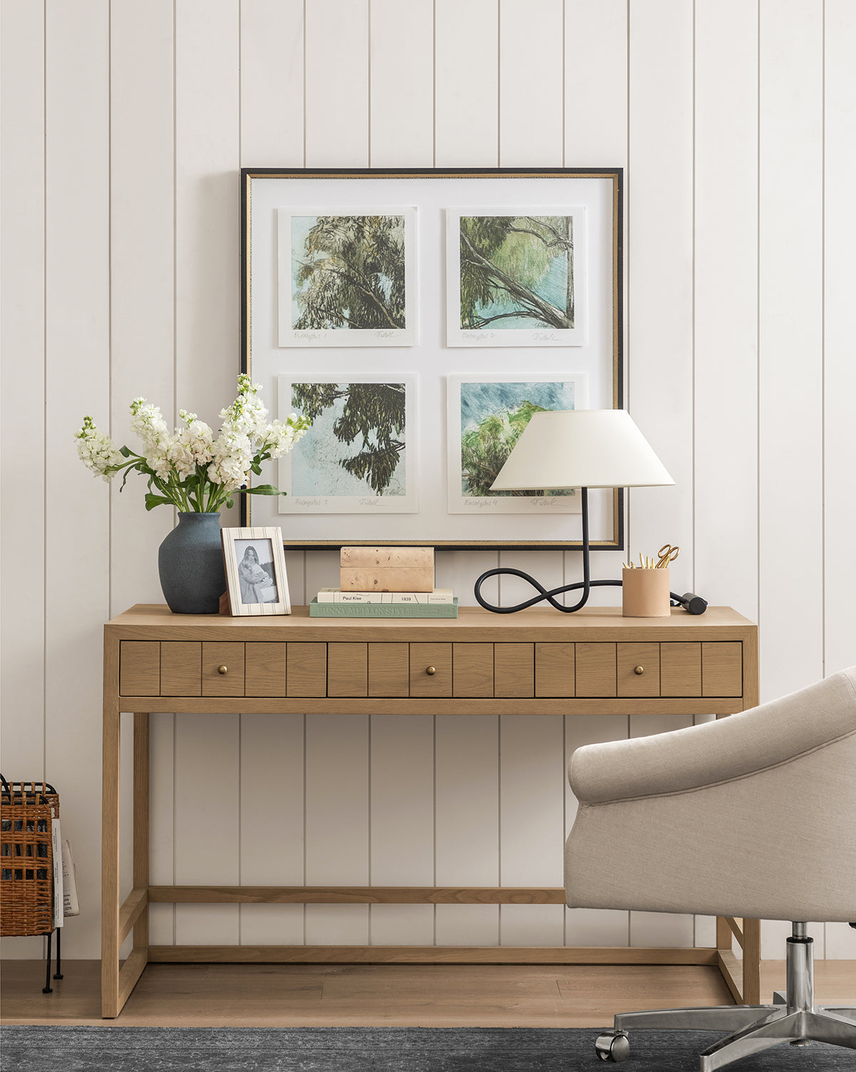 A modern wooden desk with drawers holds a lamp, white flowers, framed photo, and books. Above it, Eucalyptus Windows giclee print wall art hangs on a white shiplap wall. A light gray chair sits in front.