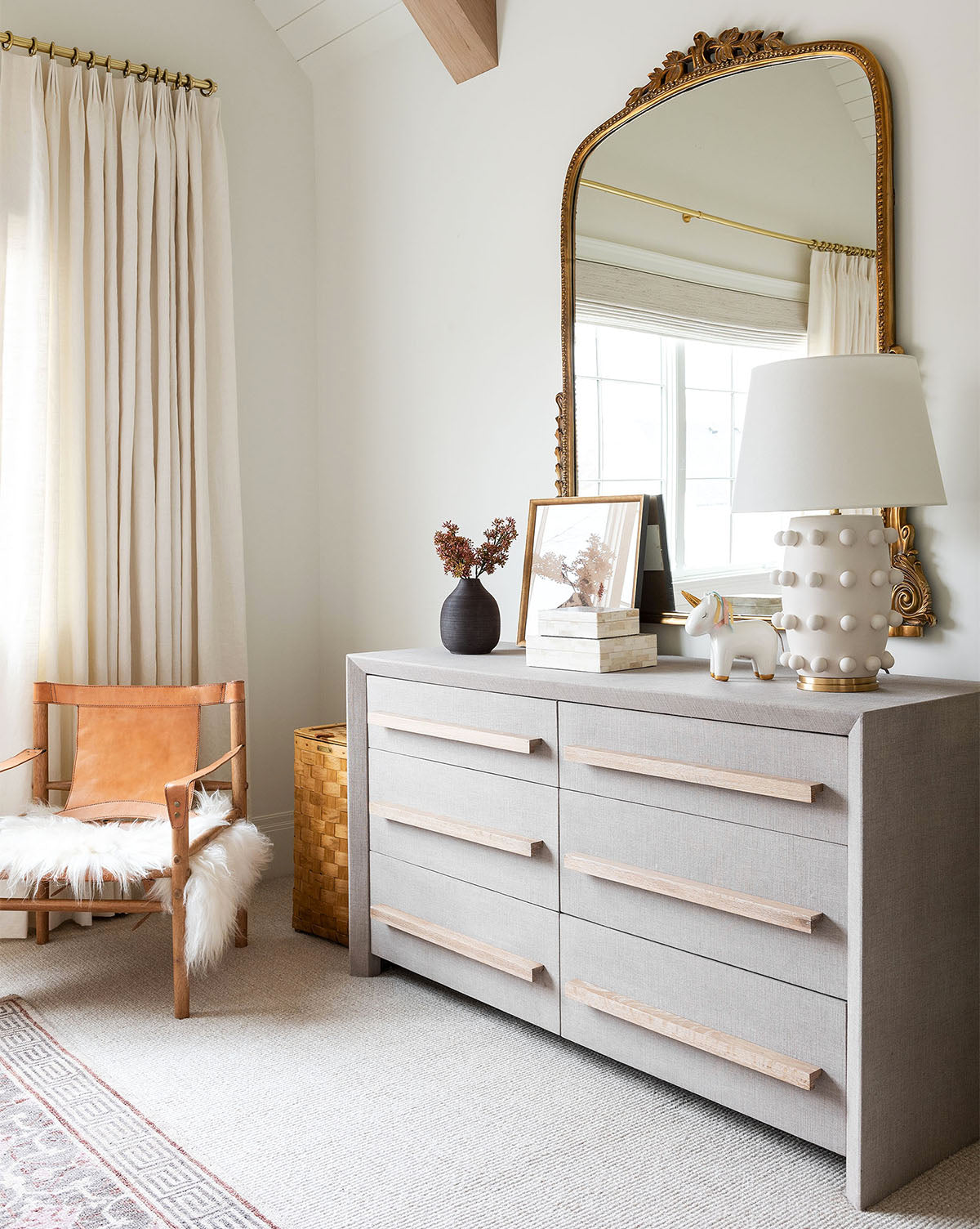 A modern bedroom corner features the Linden Table Lamp with a linen shade on a gray dresser, paired with a large ornate gold mirror, tan leather chair with white throw, and light curtains softly covering the window by McGee & Co.