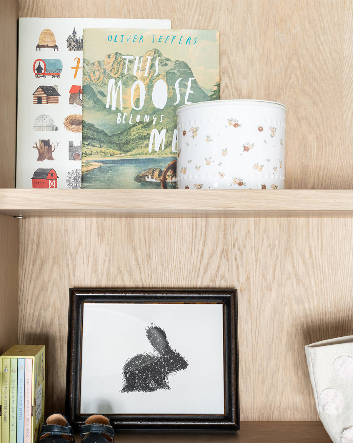A wooden shelf displays childrens picture books, a white tin with an animal pattern, and a framed black-and-white rabbit illustration. The book This Moose Belongs to Me by Oliver Jeffers is also visible.