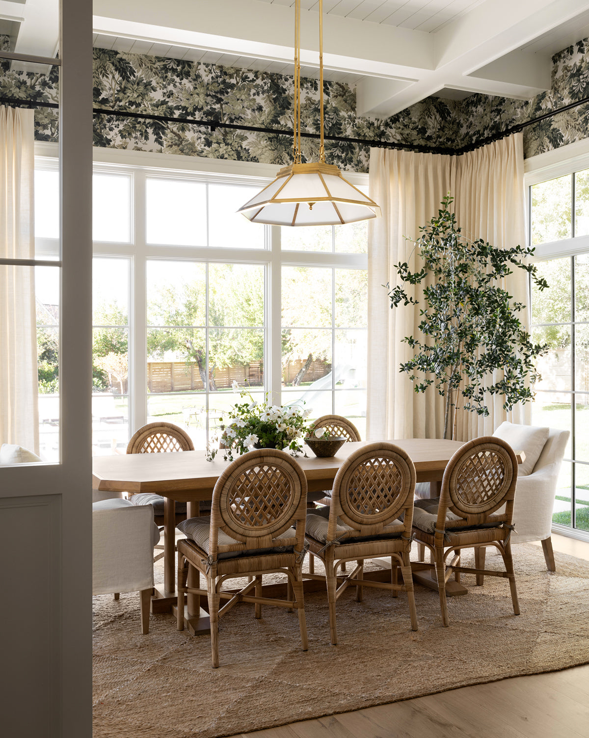 A bright dining room features the Orville Extension Dining Table, wicker chairs, and cream armchairs. Large windows fill the space with light while leafy plants and floral wallpaper create a fresh, elegant feel. A pendant light hangs above the table, by McGee & Co.
