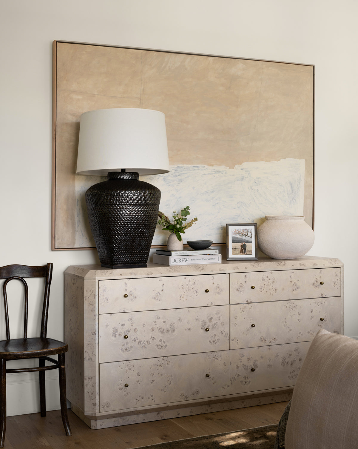 The Tali Burl Wood Dresser, topped with a dark textured lamp, books, small plant, framed photo, and round vase, sits beneath a minimalist abstract painting. A wooden chair is placed next to this modern bedroom piece by McGee & Co.