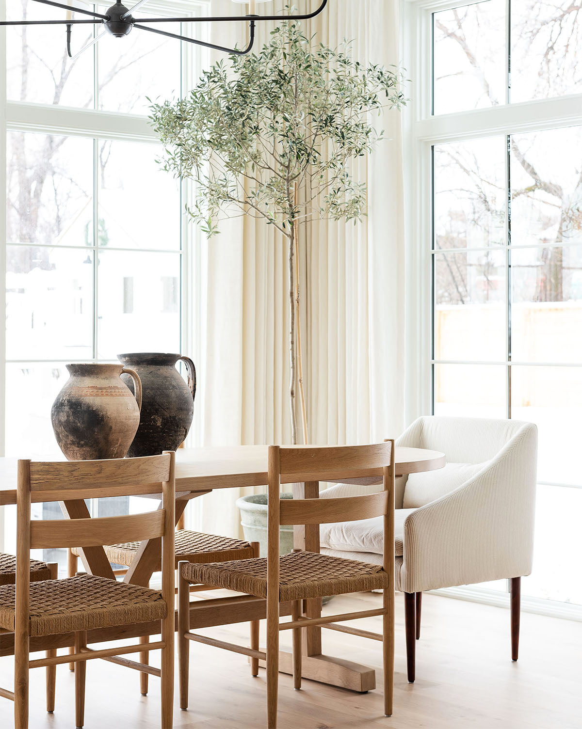 Bright dining room with Danish design touches—Eloise Woven Chairs, a white table, large pottery vases, and a tall potted tree by airy windows with sheer curtains that flood the space with natural light, by McGee & Co.