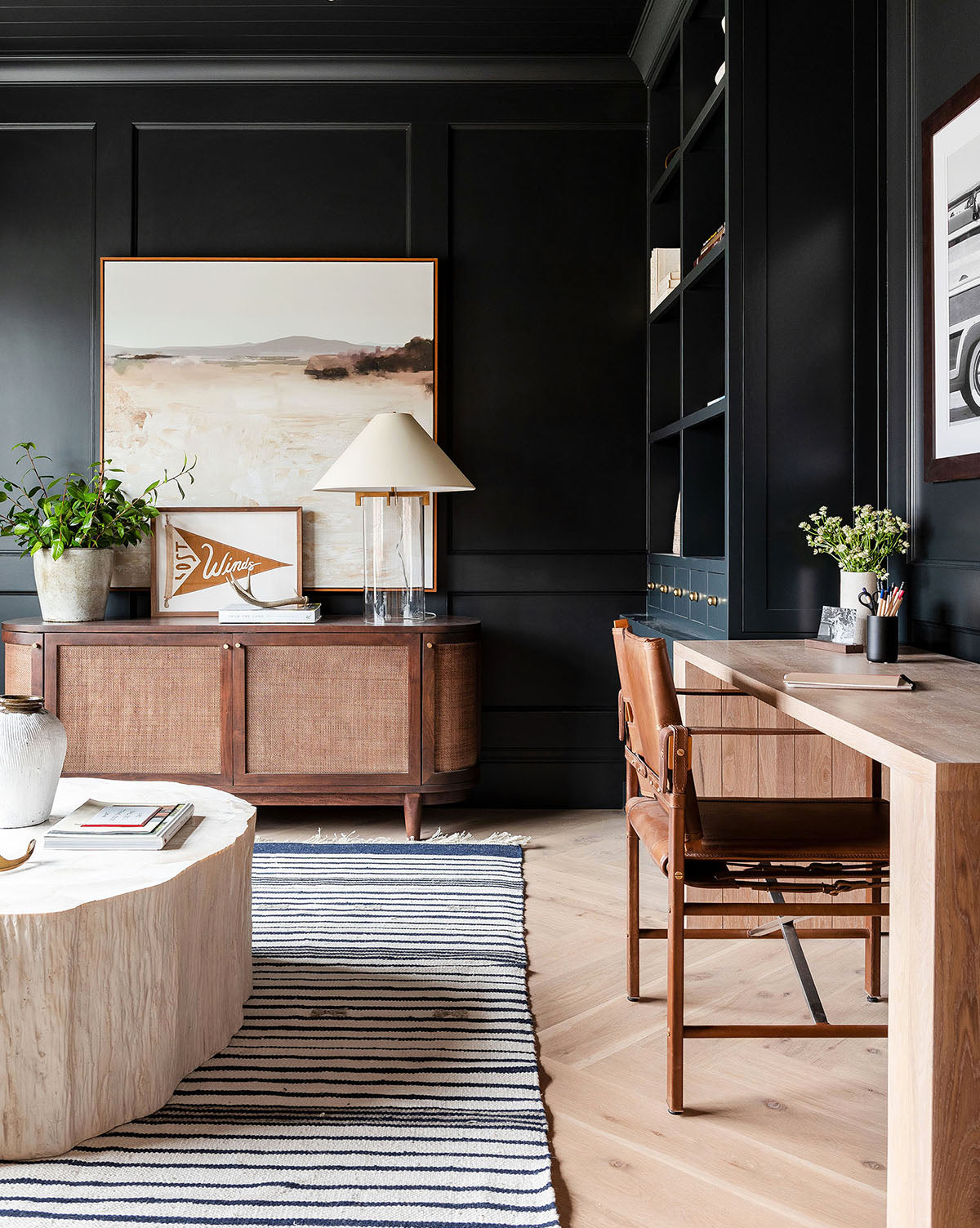 A modern home office with black walls, wooden furnishings, and a striped rug. Serene decor, plants, and the Distant Waters artwork top the sideboard, while shelves display books and relaxing art along the wall.