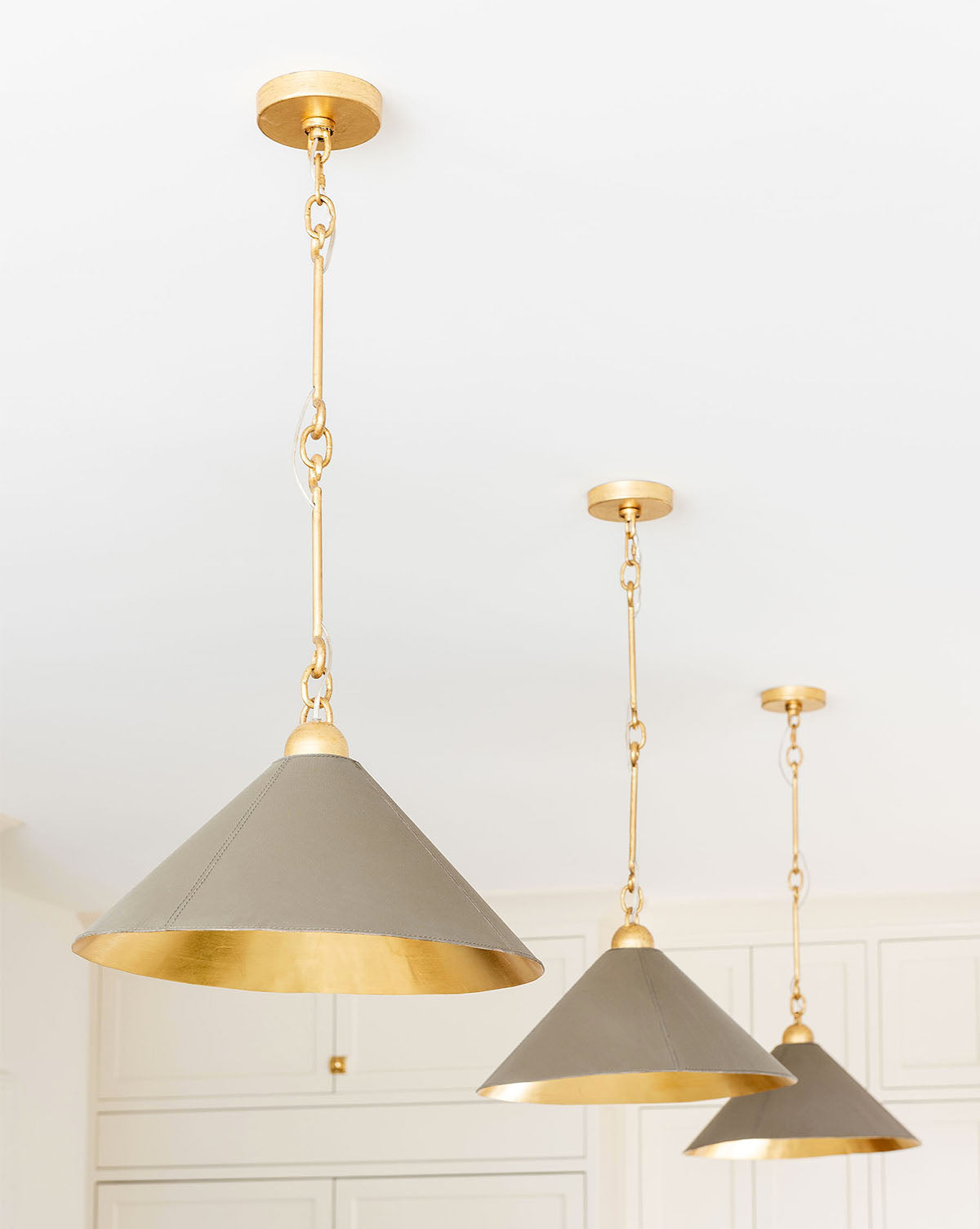 Three modern Scarlett Pendant lights hang from a white ceiling. Gold-leafed chains and fixtures support conical shades—gray outside, gold inside—creating an elegant fixture that casts a warm, sophisticated glow by McGee & Co.