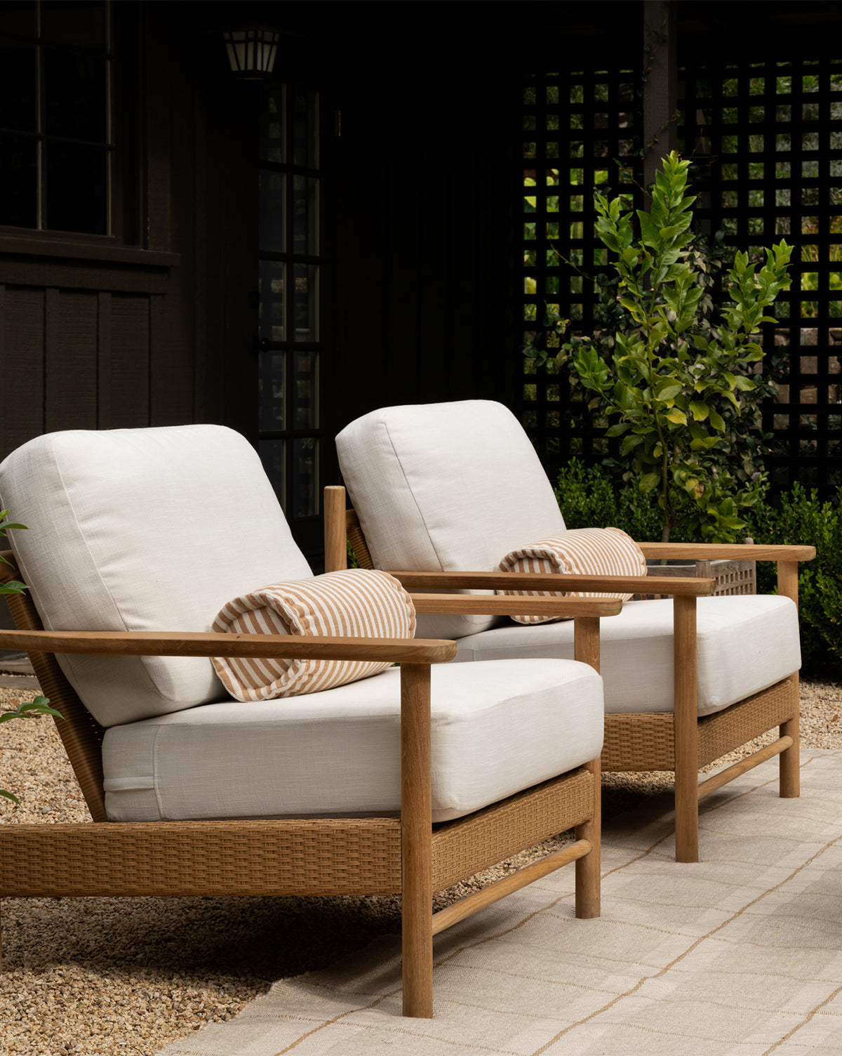 Two Simeon Outdoor Lounge Chairs with upholstered cushions and striped cylindrical pillows are placed on a stone pathway in a garden, surrounded by greenery and a dark painted fence in the background, by McGee & Co.