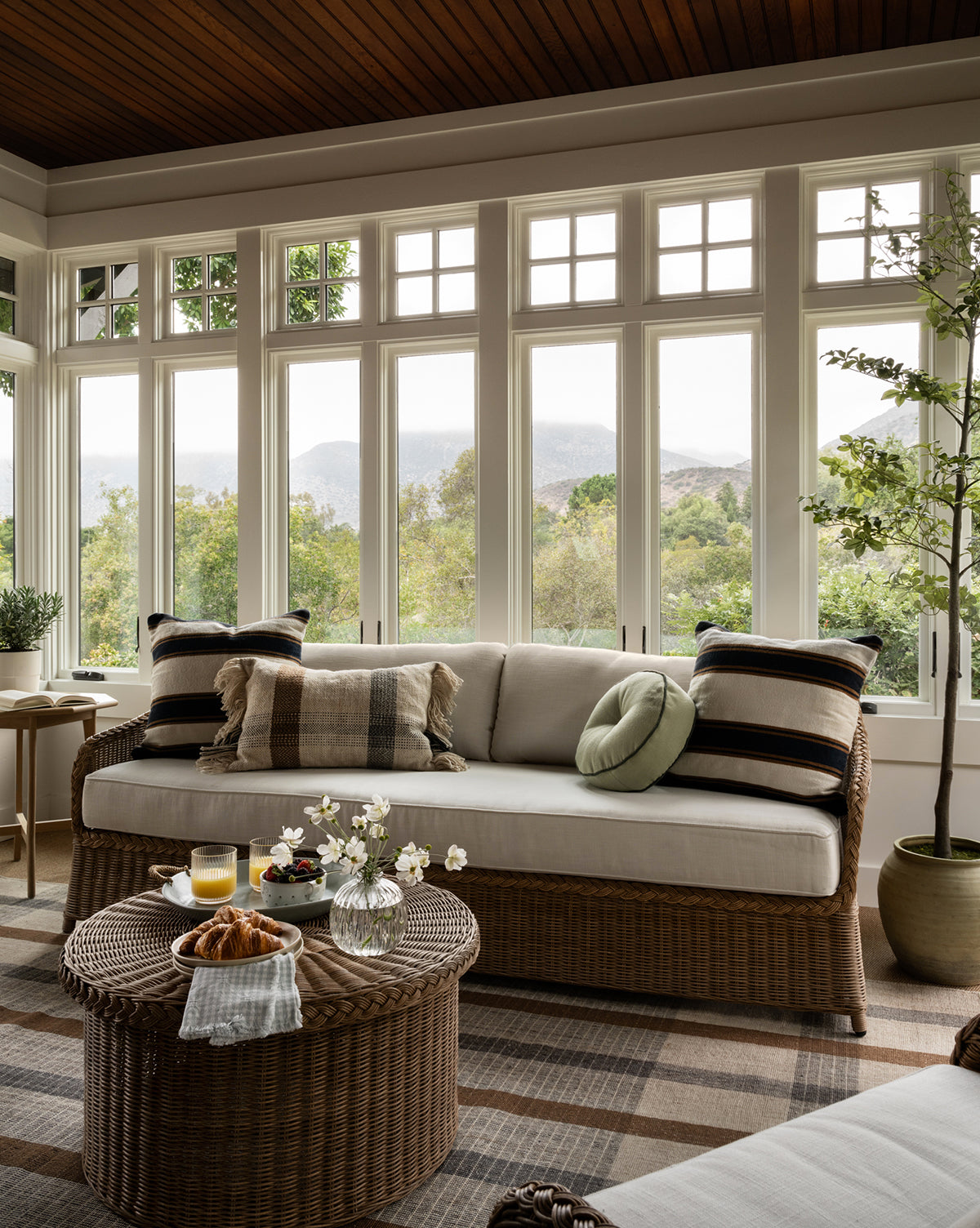 A cozy sunroom with large windows showcases the Haviland Outdoor Sofa 88 with plush pillows and a round wicker coffee table topped with drinks, flowers, and pastries. Greenery and warm natural light enhance this inviting space, by McGee & Co.