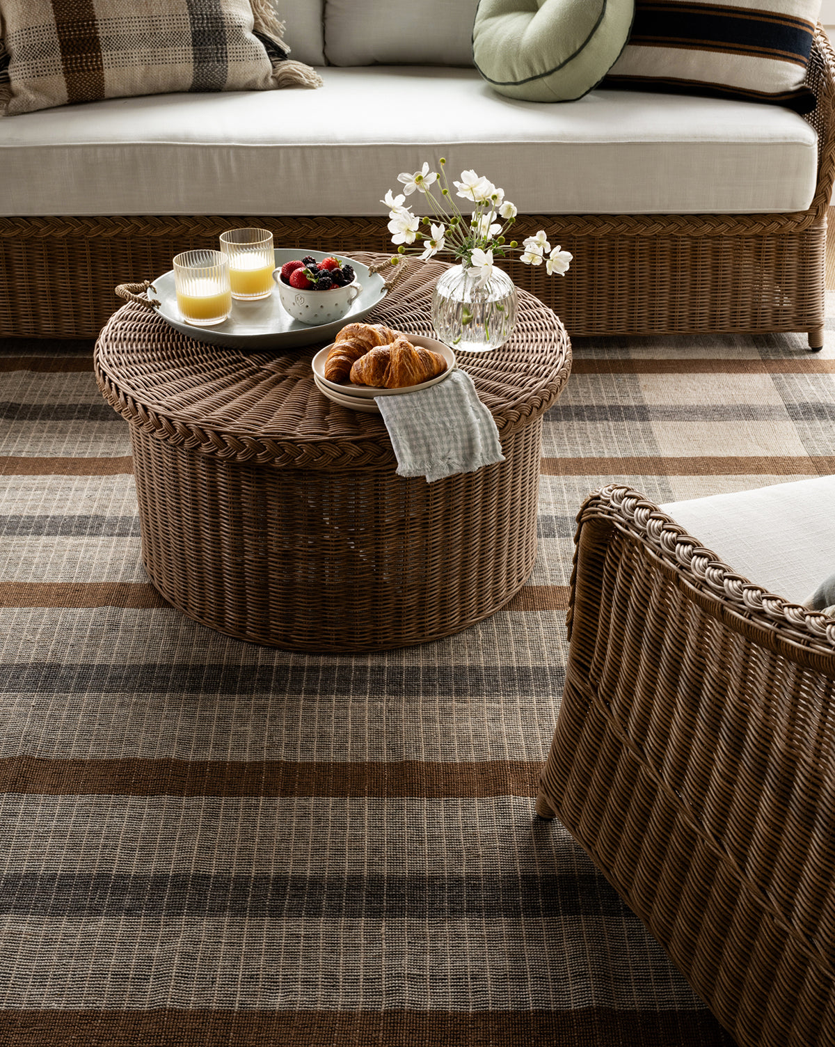 The Keenan Handwoven Indoor/Outdoor Rug lies between a wicker sofa and chair with cream cushions, topped by a wicker coffee table holding croissants, juice, berries, and flowers in a cozy, sunlit room by McGee & Co.