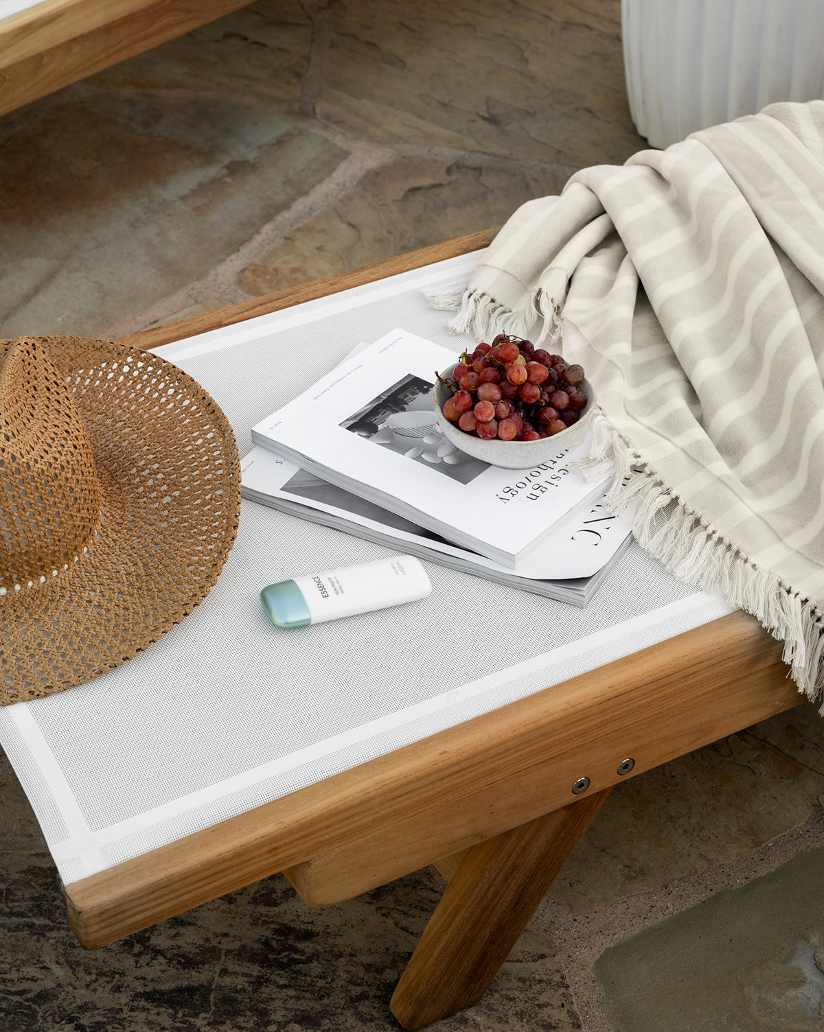 The Cadie Outdoor Chaise holds a striped blanket, straw hat, white skincare tube, bowl of grapes, and magazines, all arranged on a stone patio by McGee & Co.