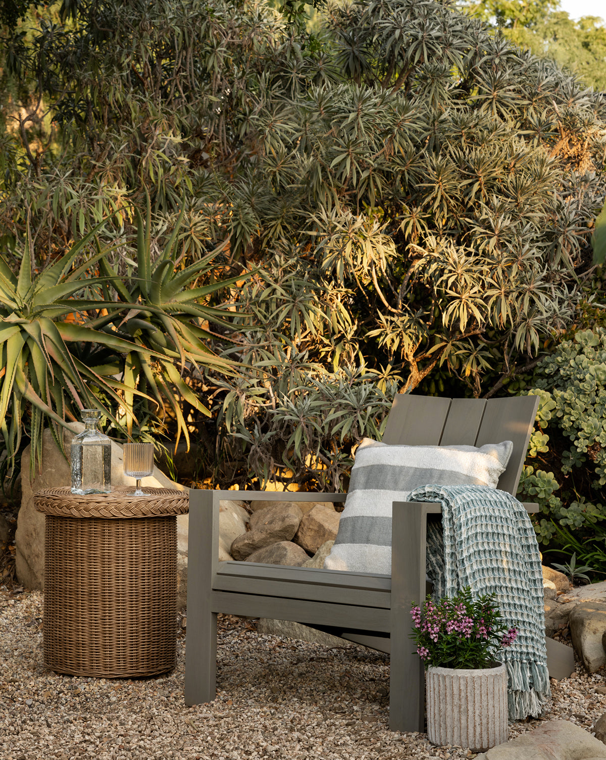The Triby Gray Teak Outdoor Lounge Chair with a striped cushion and blue blanket sits on gravel beside a wicker side table with glassware, surrounded by lush greenery and potted flowers in a tranquil garden setting by McGee & Co.