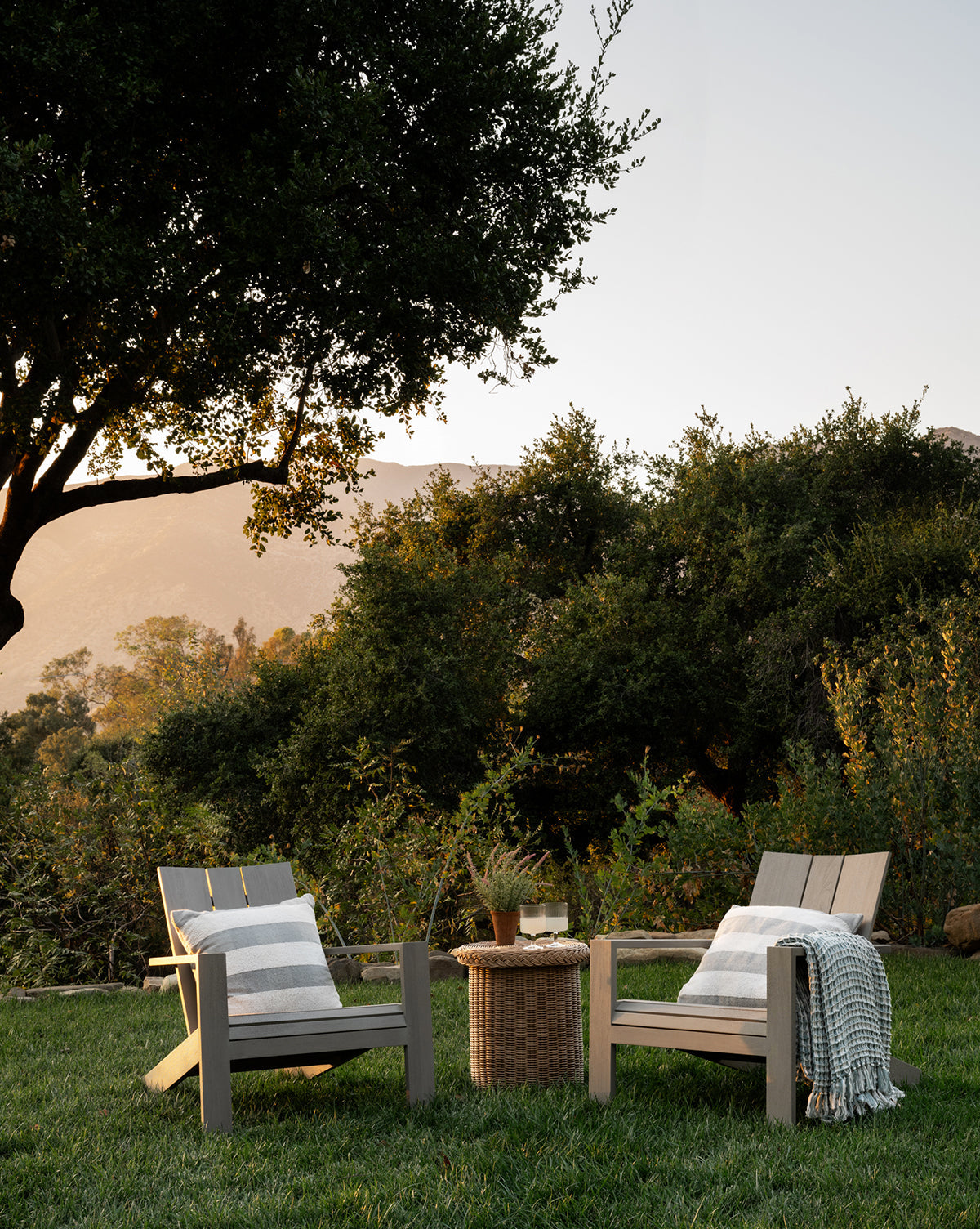 Two Triby Gray Teak Outdoor Lounge Chairs with cushions and throws sit on grass facing a small round wicker table with drinks and a plant, surrounded by greenery and trees, with mountains visible in the sunset—a relaxing outdoor setup, by McGee & Co.