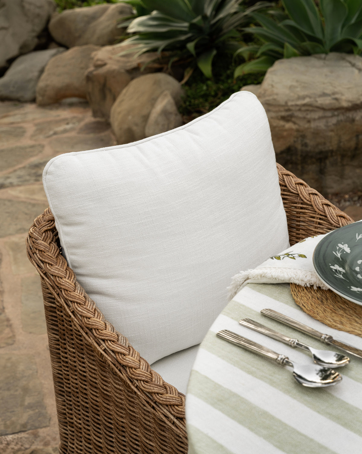 The Haviland Outdoor Dining Chair with a white cushion sits by a round table draped in a green and white striped cloth, ready for outdoor dining. Surrounded by rocks and plants, it creates an inviting backyard setting by McGee & Co.
