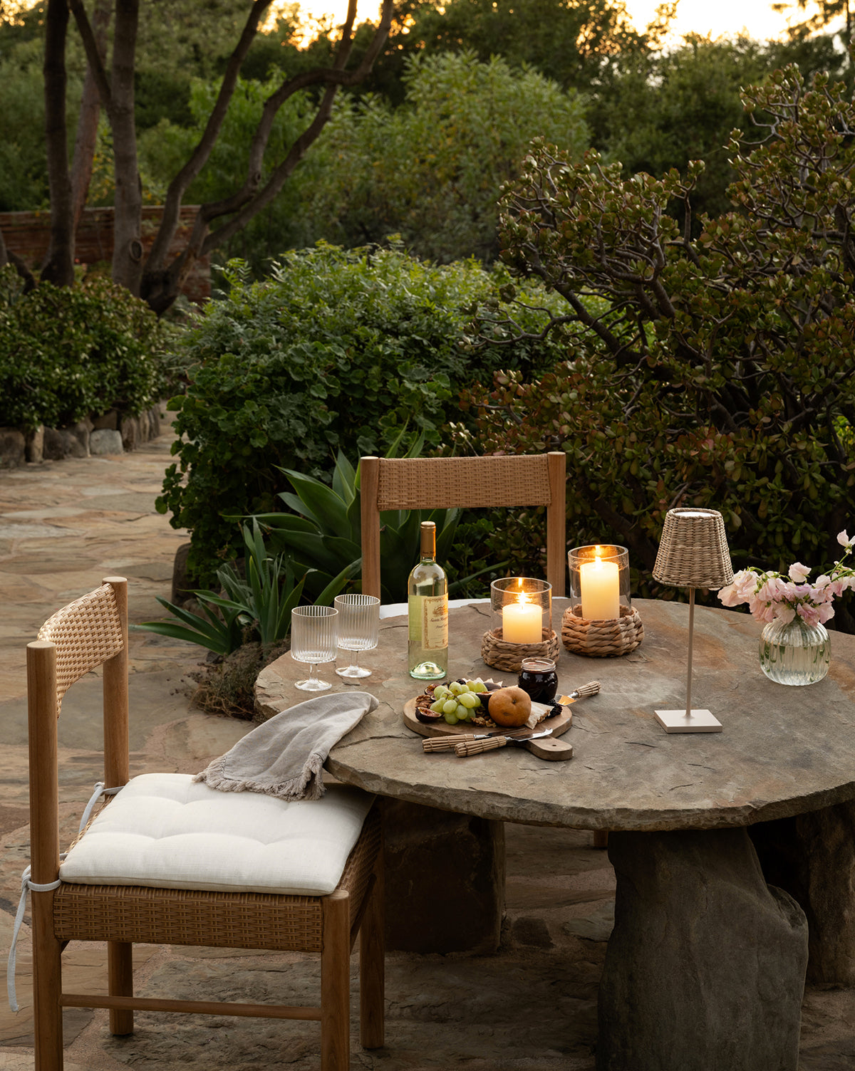 A rustic outdoor table with a Simeon Outdoor Dining Chair, another chair, candles, wine, glasses, snacks, and flowers sits in a lush garden at dusk. Greenery and stone paving surround the inviting scene by McGee & Co.