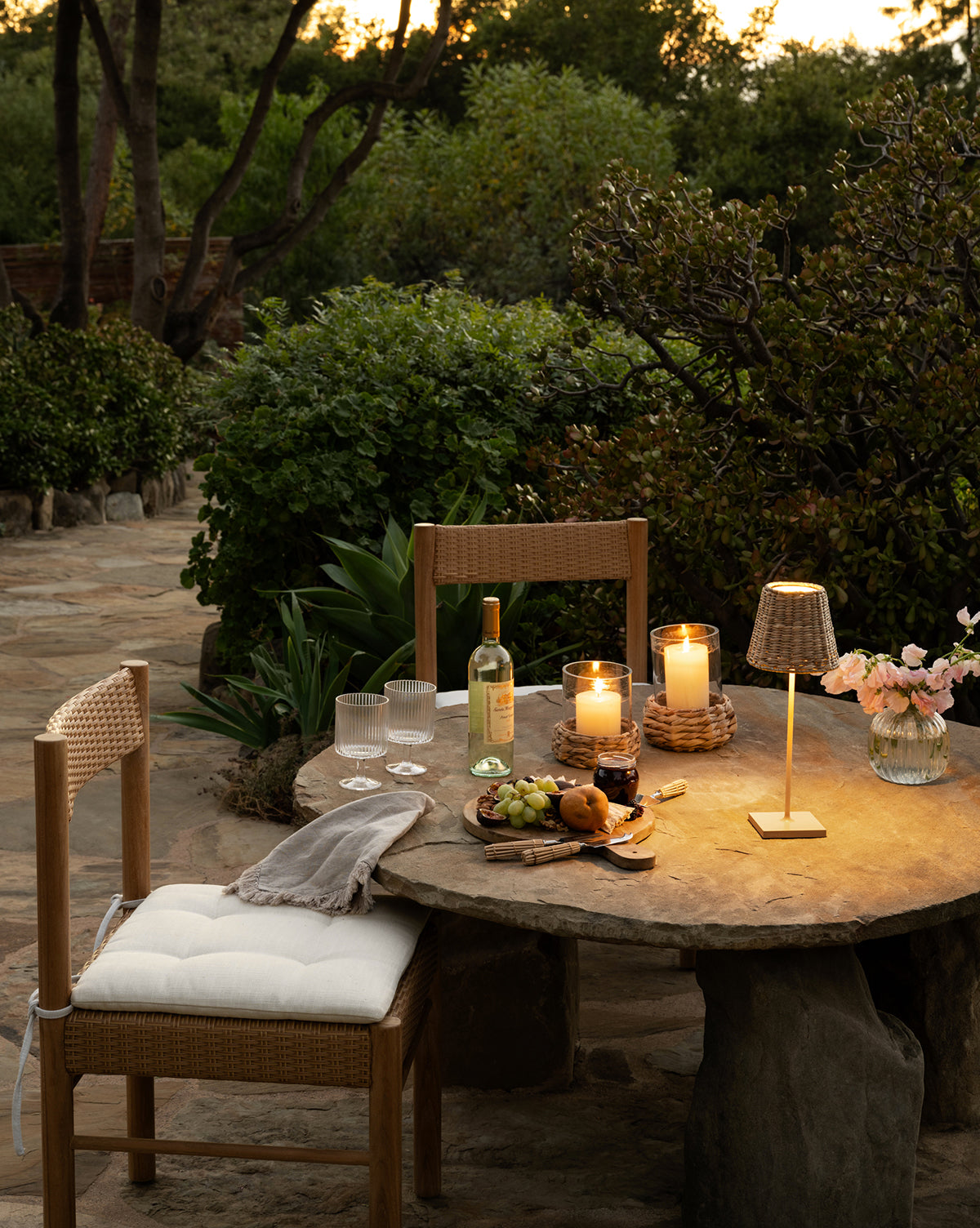 A stone patio table holds two wooden chairs, lit candles, a Seagrass Lamp Shade, white wine with two glasses, a fruit plate, and a vase of flowers—all surrounded by lush sunset greenery, by McGee & Co.
