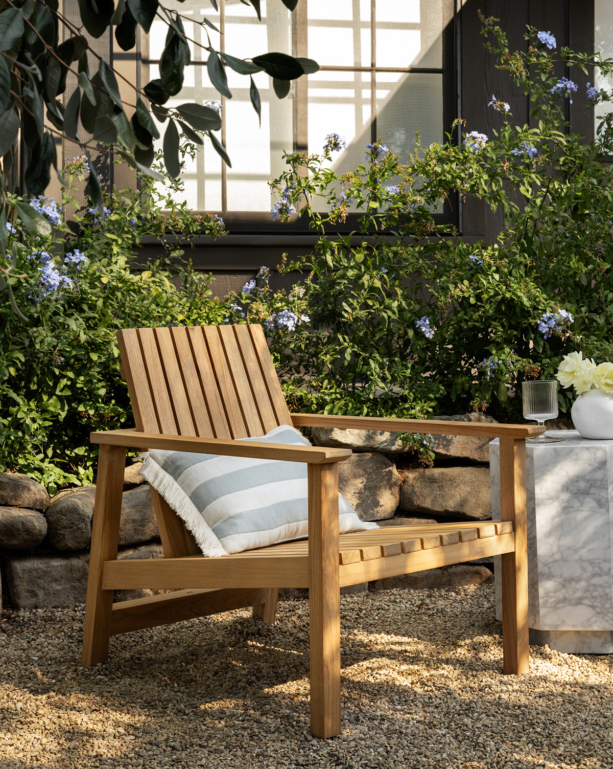 The Gilda Outdoor Lounge Chair with a striped cushion rests on gravel beside a small marble side table with flowers, set against lush green bushes and blooming purple flowers in front of a house’s window, by McGee & Co.
