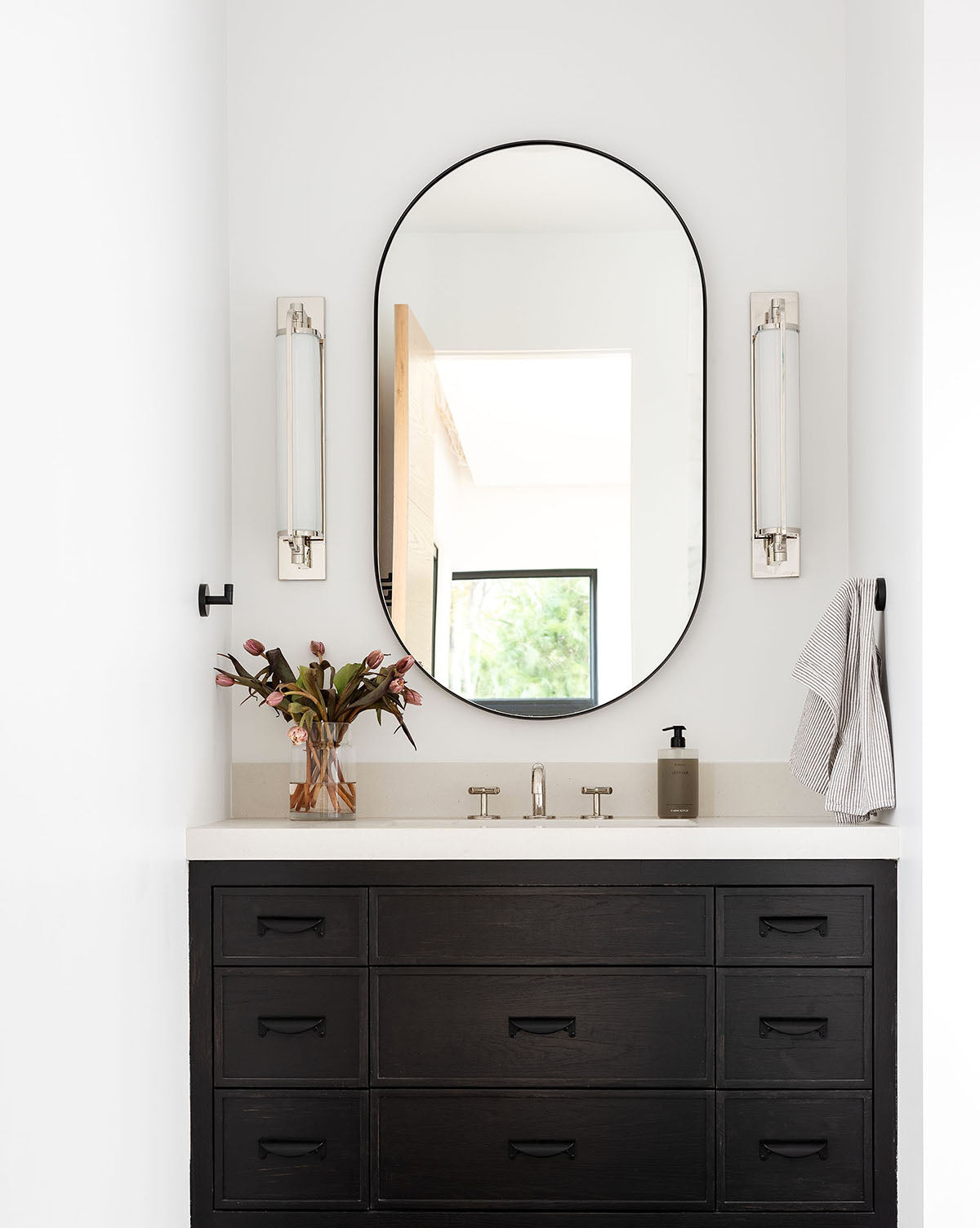 A modern bathroom vanity features a black cabinet, white countertop, Rye Wall Mirror, wall-mounted lights, vase of flowers, stainless steel soap dispenser, and a striped towel hanging on the right wall, by McGee & Co.