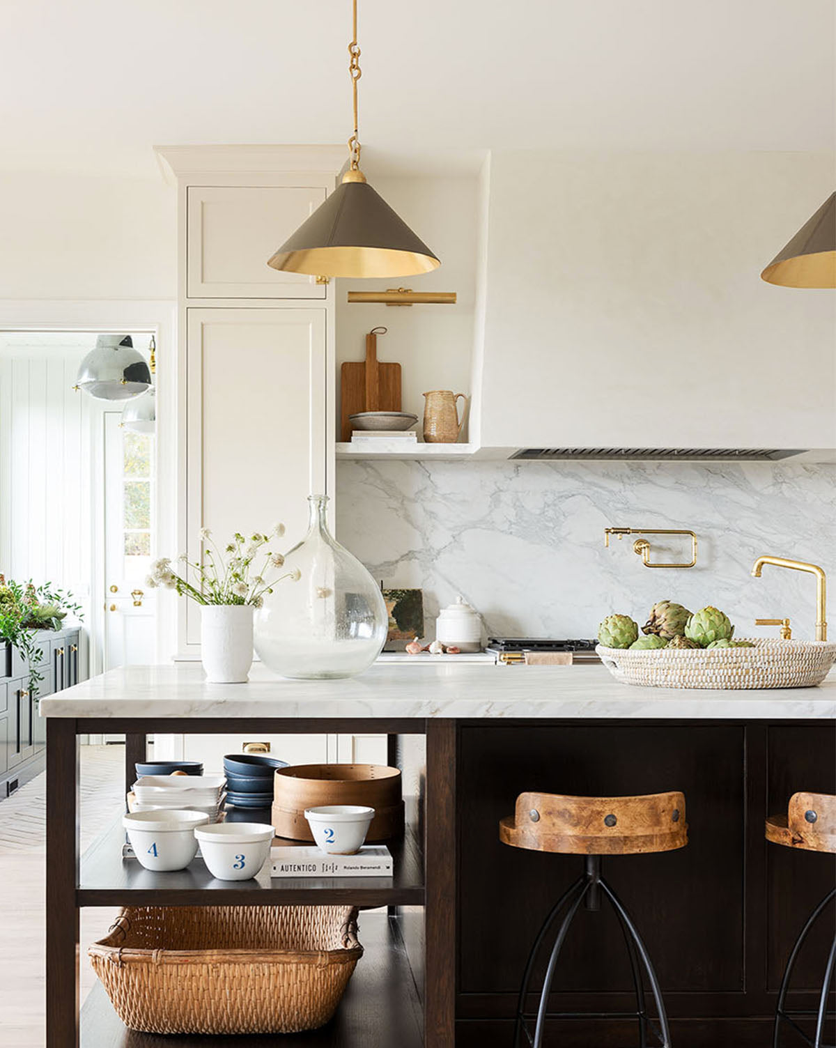 A bright, modern kitchen with a marble backsplash, gold fixtures, a dark wood island with open shelves, two wooden stools, and elegant Scarlett Pendants illuminating the counter by McGee & Co.