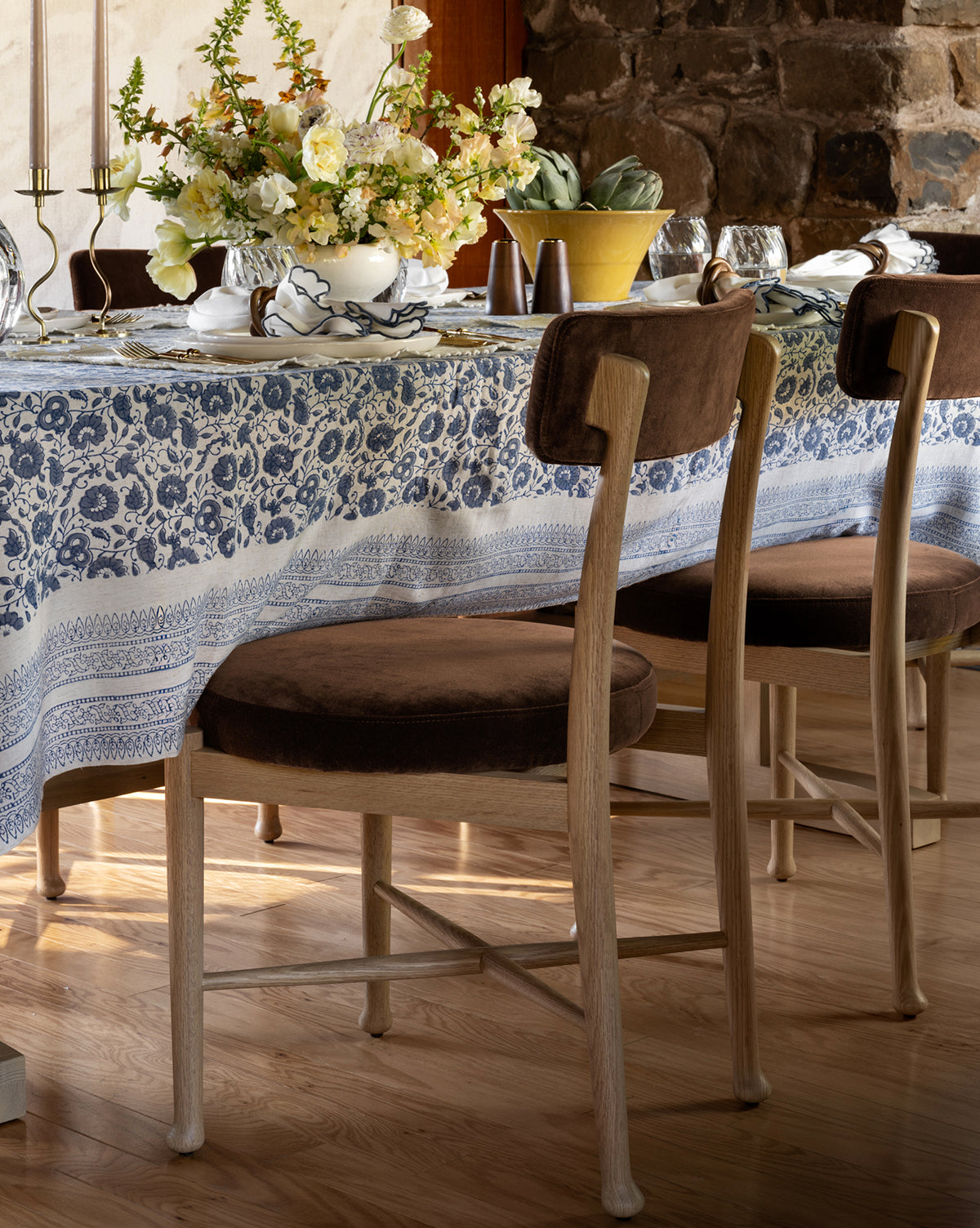 A dining table with a blue and white patterned tablecloth, floral centerpieces, dishes, and candlesticks is set on a wooden floor. Two Garance Dining Chairs with brown cushions are positioned beside the table by McGee & Co.