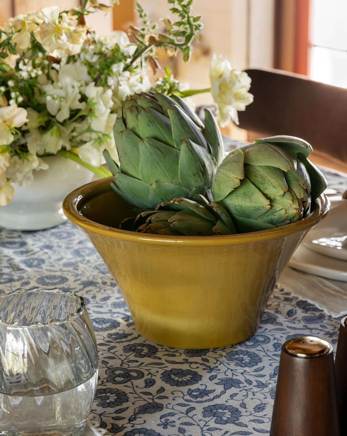 The Brennan Serving Bowl, filled with two artichokes, rests on a patterned tablecloth beside a glass of water, a white vase of flowers, and a brown salt shaker. Sunlight from the window brightens the kitchen decor, by McGee & Co.