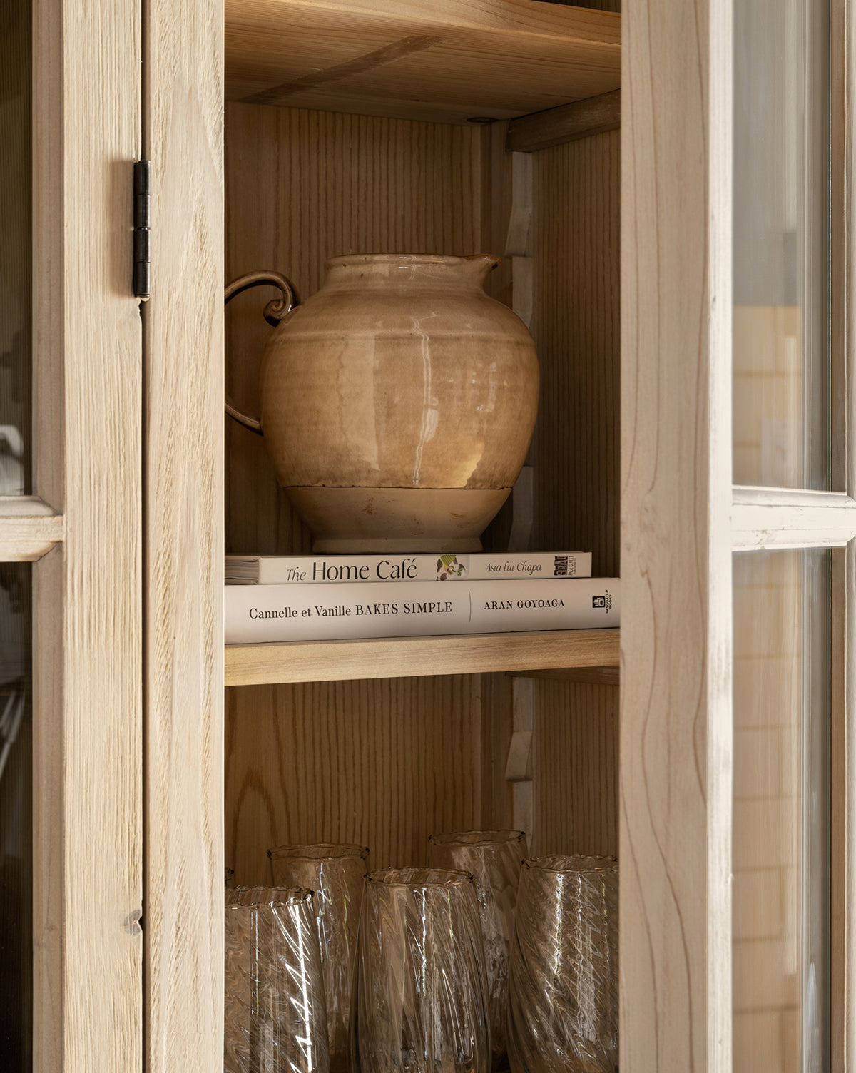 A Brown Stoneware Pitcher sits atop two stacked cookbooks inside a light wood cabinet, while several textured glass tumblers are neatly arranged below—adding style to any kitchen, by McGee & Co.
