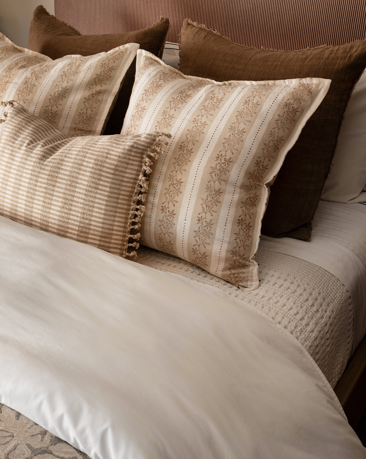 A neatly made bed with large brown floral pillows, solid brown pillows, a small beige checked tassel pillow, all set against a textured headboard and cream bedding topped with the 100% cotton Portuguese Carnel Waffle Blanket.