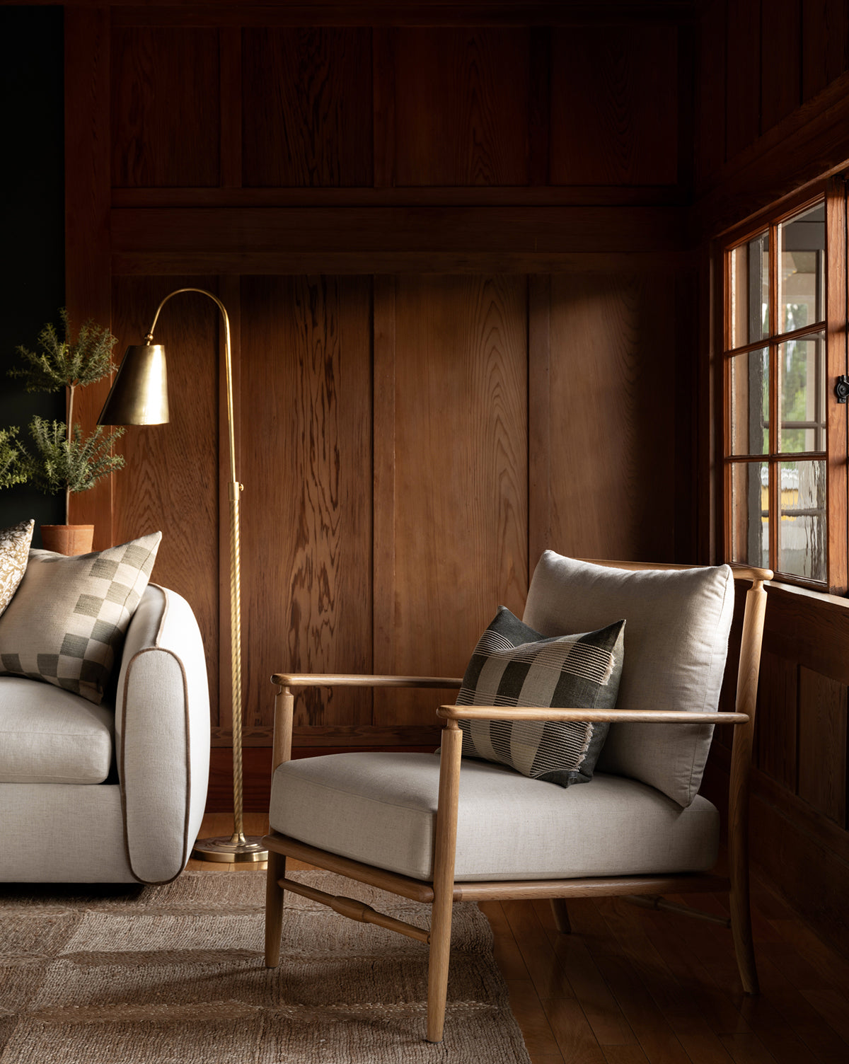 A cozy living room with wood-paneled walls, a Fenwick Lounge Chair, matching sofa, patterned pillows, brass floor lamp, and sunlight pouring through a window onto the rug, by McGee & Co.