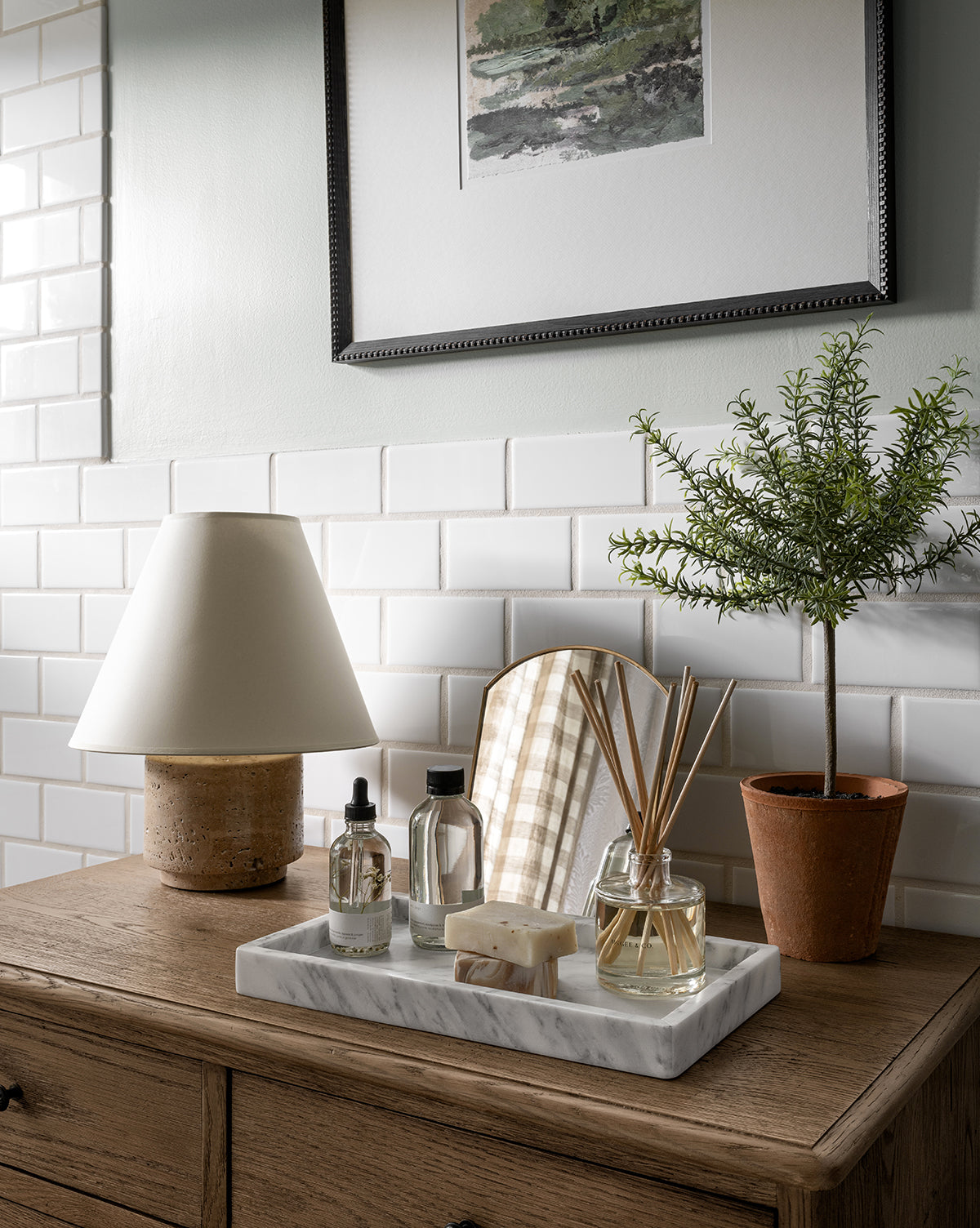 A standing vanity mirror sits on a wooden dresser with a lamp, potted plant, and marble tray holding bottles, soap, and a reed diffuser. Brass-finished metal frame and white subway tiles accent the bathroom countertop decor with framed artwork behind.