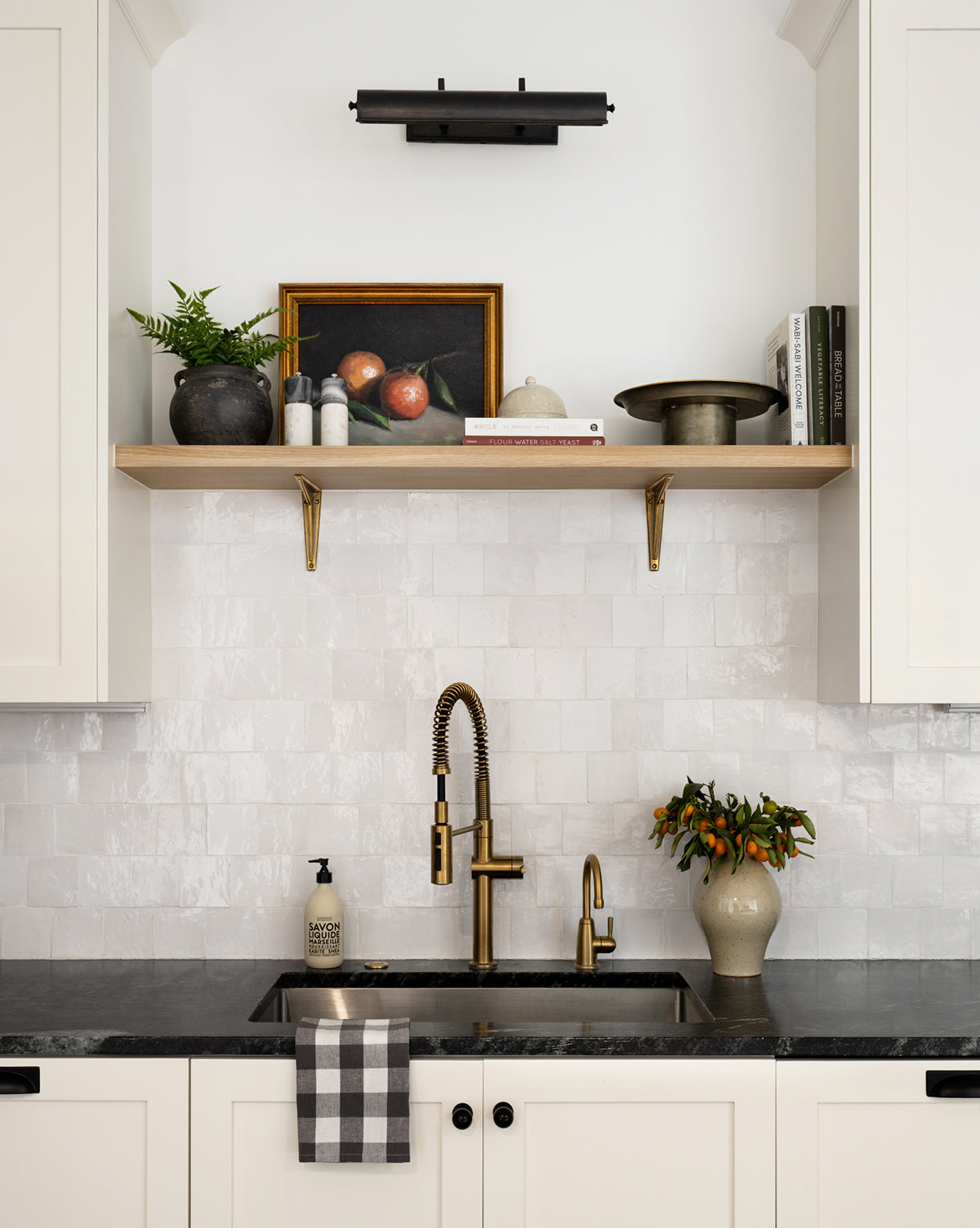 A modern kitchen sink with a black countertop, brass faucet, checkered towel, and a vase with orange berries. Above, a wood shelf displays the product Still Life with Oranges art, books, plants, and decor on a white tile backsplash.
