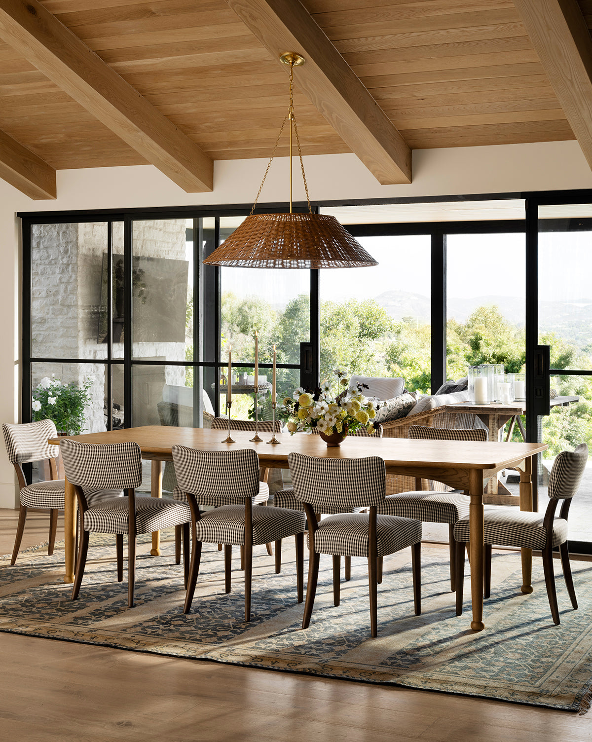 A dining room featuring the handcrafted Phipps Dining Table, eight upholstered chairs, a woven pendant light, wood-beam ceilings, a patterned rug, and large glass doors opening to a patio with greenery by McGee & Co.