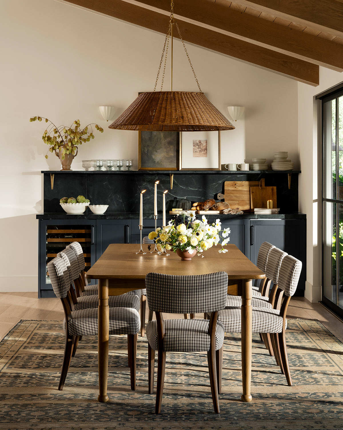 A cozy dining room with the handcrafted Phipps Dining Table, six checkered chairs, a floral centerpiece, and lit candles. Dark cabinets, open shelves with dishes, and a woven pendant light complete the modern rustic décor by McGee & Co.