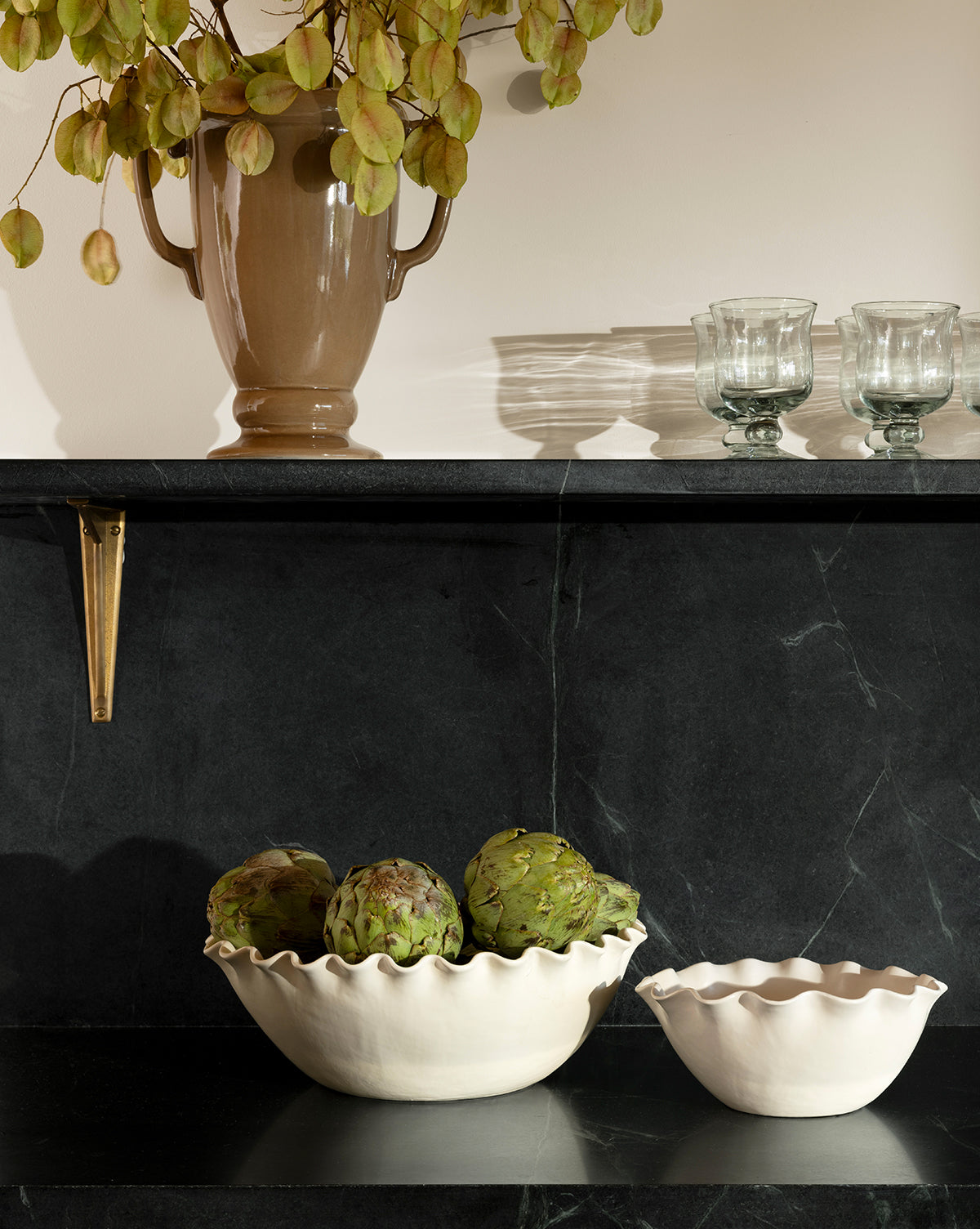 A dark marble shelf displays the Olindo Serving Bowls (Set of 2), one filled with artichokes. Above, a beige vase with dried leaves and two clear glasses are arranged against a pale wall, by McGee & Co.