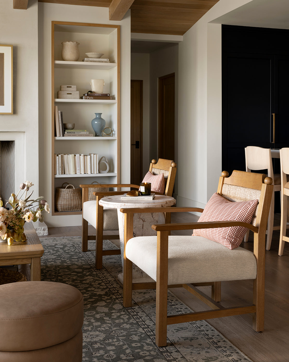 A cozy living room features the Alberta Lounge Chair, a patterned rug, and a round ottoman. Shelves display books and ceramics, while neutral tones and soft lighting give the space a warm, inviting feel by McGee & Co.