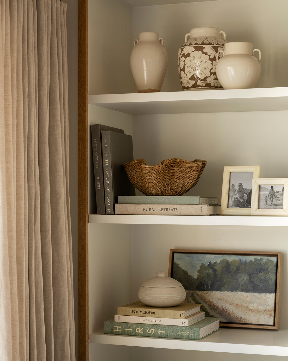 A bookshelf with stacked books, the Goulding Woven Bowl, beige ceramic vases, framed photos, and a landscape painting beside beige curtains. Decor features neutral and earthy tones, by McGee & Co.