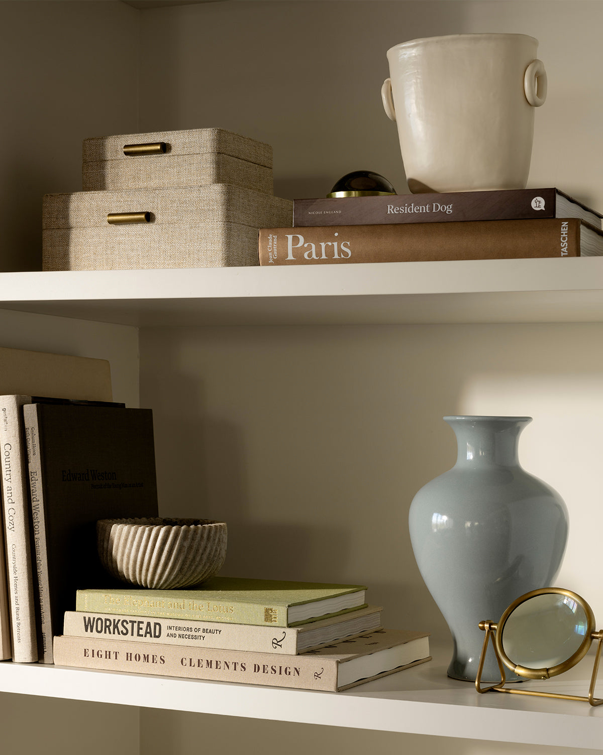 Two white shelves display neutral-toned books, a pale blue vase, a Beige Spiral Marble Bowl, stacked linen boxes, a cream ceramic pot, a gold magnifying glass, and various decorative objects in an organized arrangement by McGee & Co.