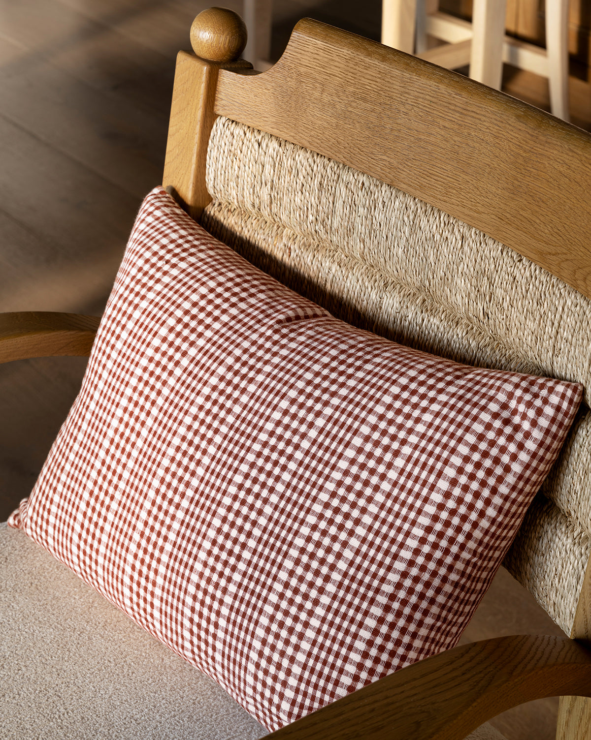 A wooden chair with a woven backrest and seat is accented by the Spencer Gingham Pillow Cover in red and white, illuminated by natural light, by McGee & Co.