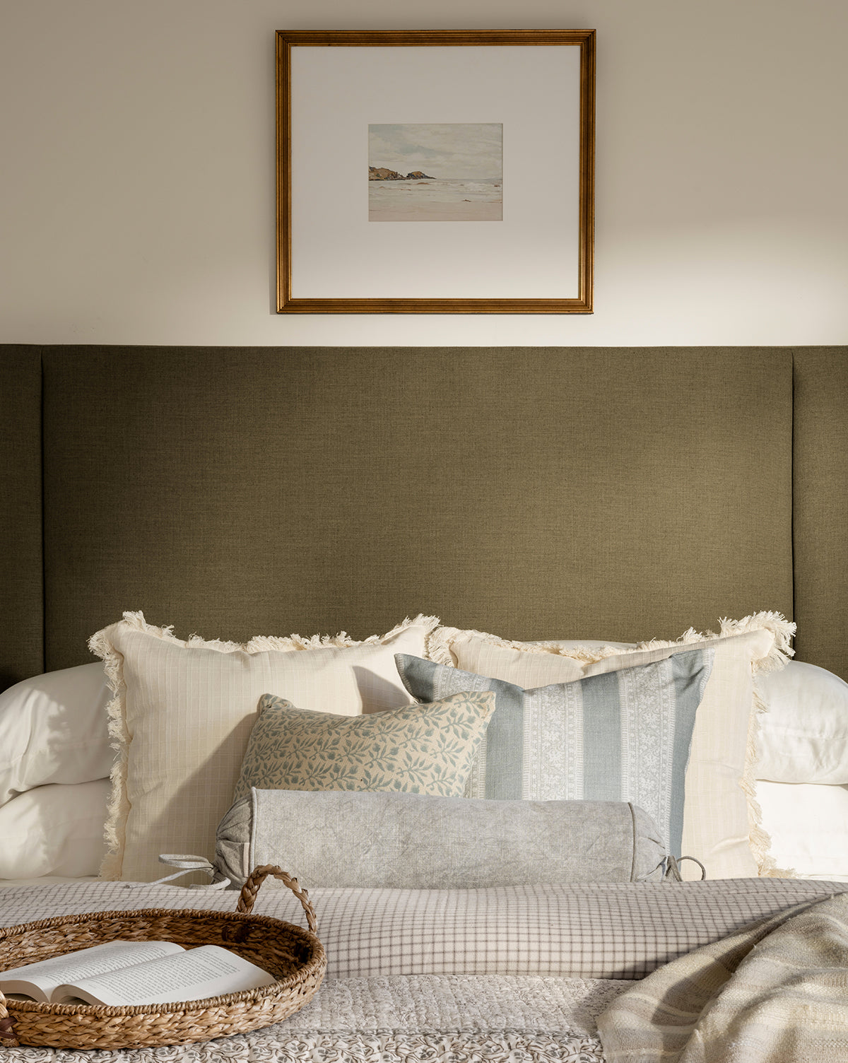 A neatly made bed with decorative pillows, including the Delphine Bolster Pillow, a woven tray at the foot, and a neutral fabric headboard. A framed landscape artwork hangs above the headboard on a beige wall.