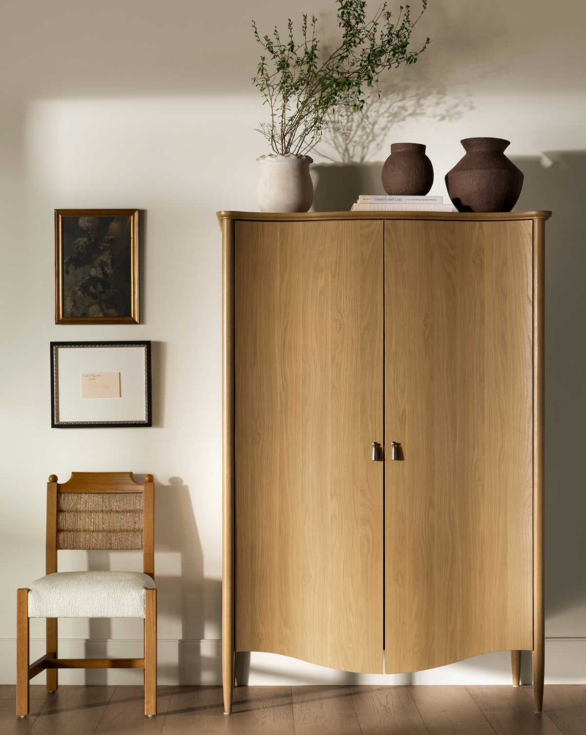 A Moorland Cabinet with books, two brown vases, and a plant on top stands by a wooden chair with a woven seat. Three framed artworks hang above the chair, adding stylish storage to the living rooms wood floor, by McGee & Co.