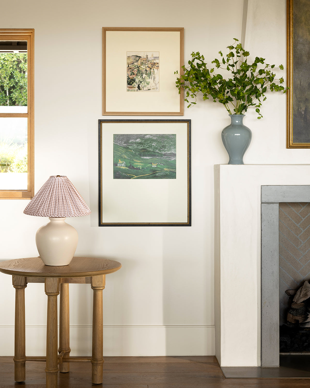 A cozy interior features the Country Dusk giclee prints in black and gold frames above a mantel with a light blue vase of green branches, plus a small round wooden table with a ceramic lamp. A window reveals greenery outside, by McGee & Co.