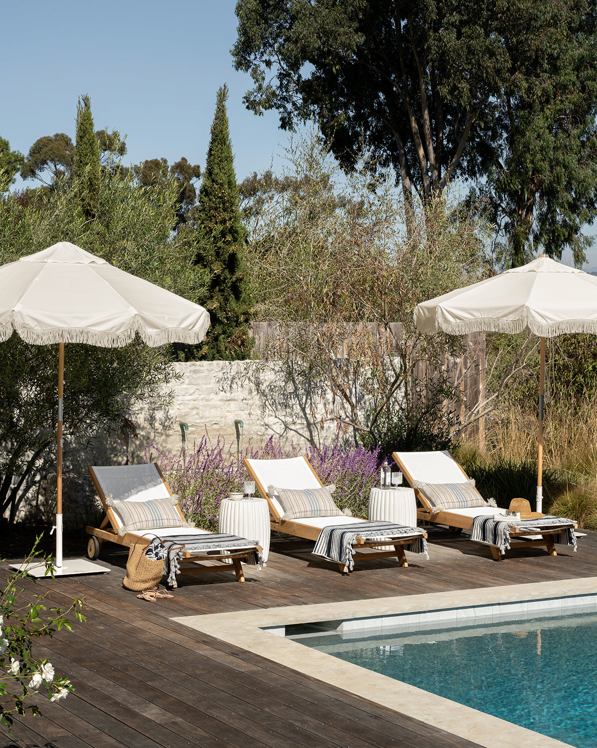 Three Cadie Outdoor Chaises with striped, weather-resistant cushions are arranged by the pool on a wooden deck under two large white umbrellas, surrounded by lush trees and greenery for a tranquil ambiance, by McGee & Co.