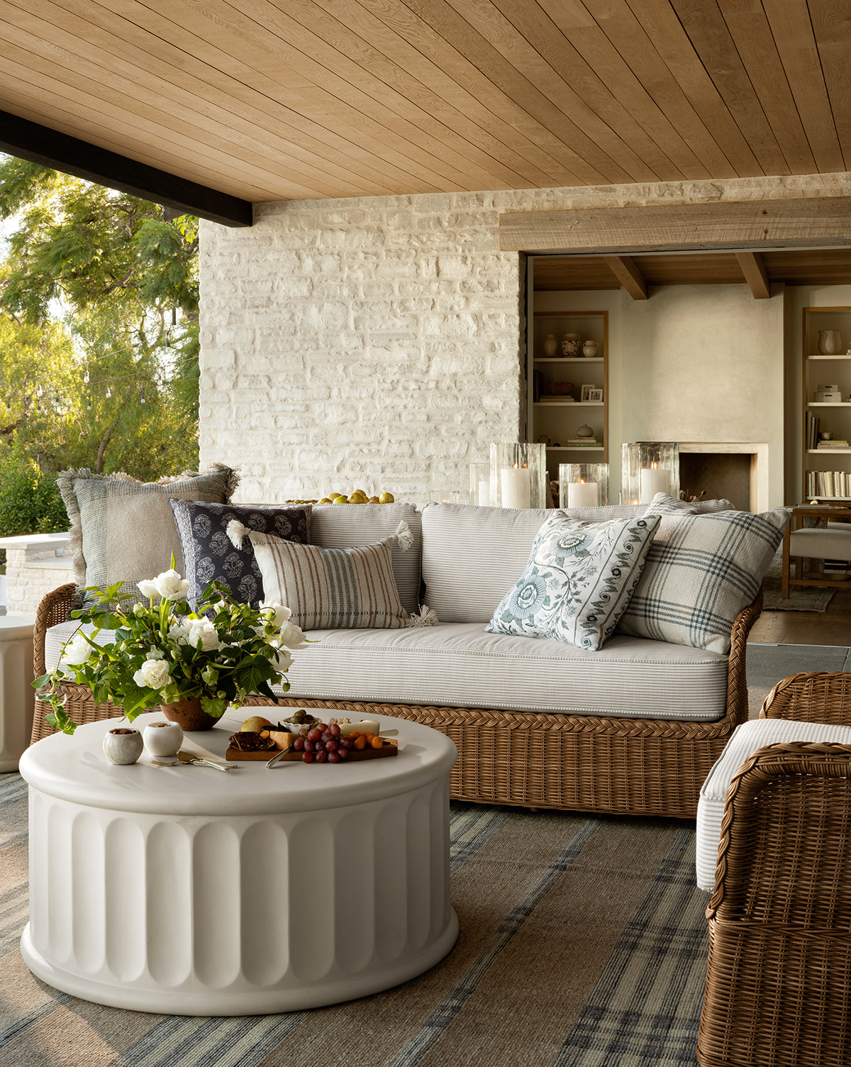 A cozy outdoor patio features the Haviland Outdoor Sofa with Perennials Taupe Stripe 88, patterned cushions, a white round coffee table with flowers and snacks, a rug over wooden flooring, and greenery visible outside by McGee & Co.