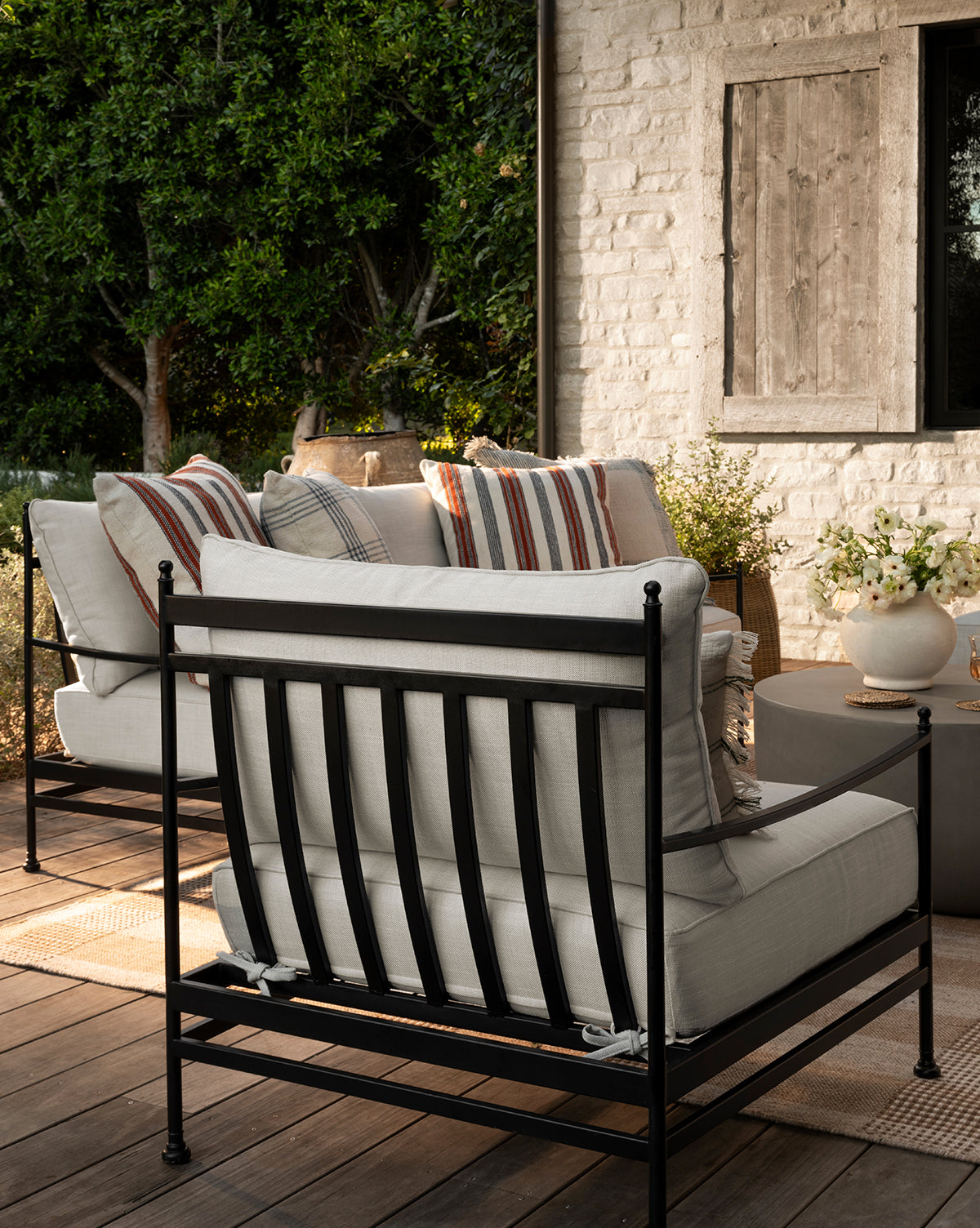 A cozy patio showcases Pomeroy Outdoor Lounge Chairs with striped pillows in outdoor fabric, a round coffee table with flowers, and a white brick wall with wood shutter, all amid lush greenery by McGee & Co.