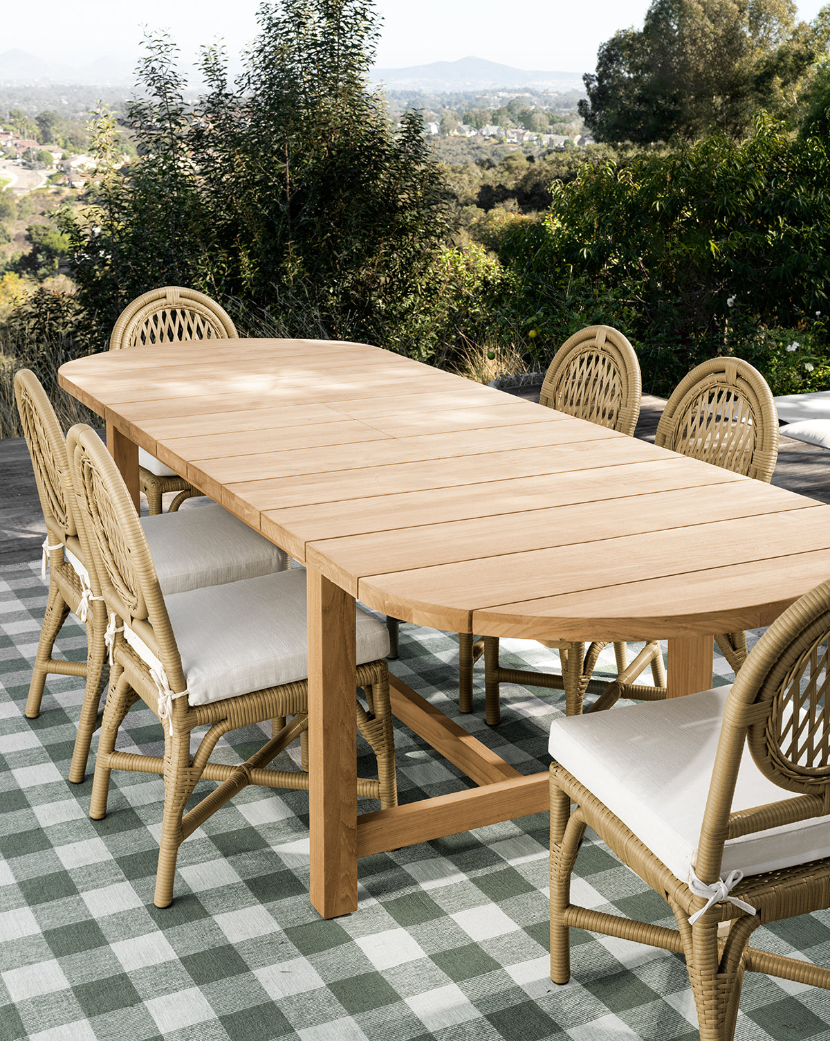 The Linwood Teak Extension Dining Table with eight wicker chairs sits on a green and white checkered rug on the patio, surrounded by lush trees and distant hills, by McGee & Co.