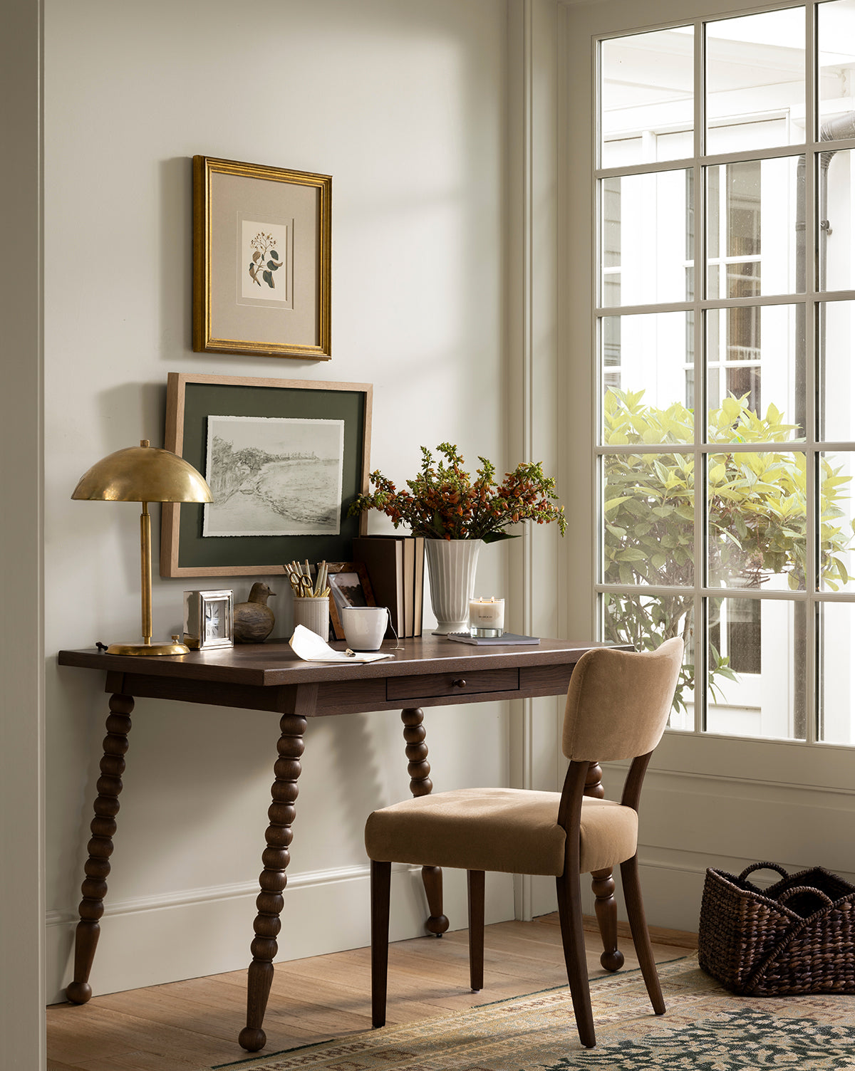 A small home office nook with a Heathcliff Desk, beige upholstered chair, gold lamp, framed art, vases of flowers, and a window bringing in natural light with views of greenery outside, by McGee & Co.
