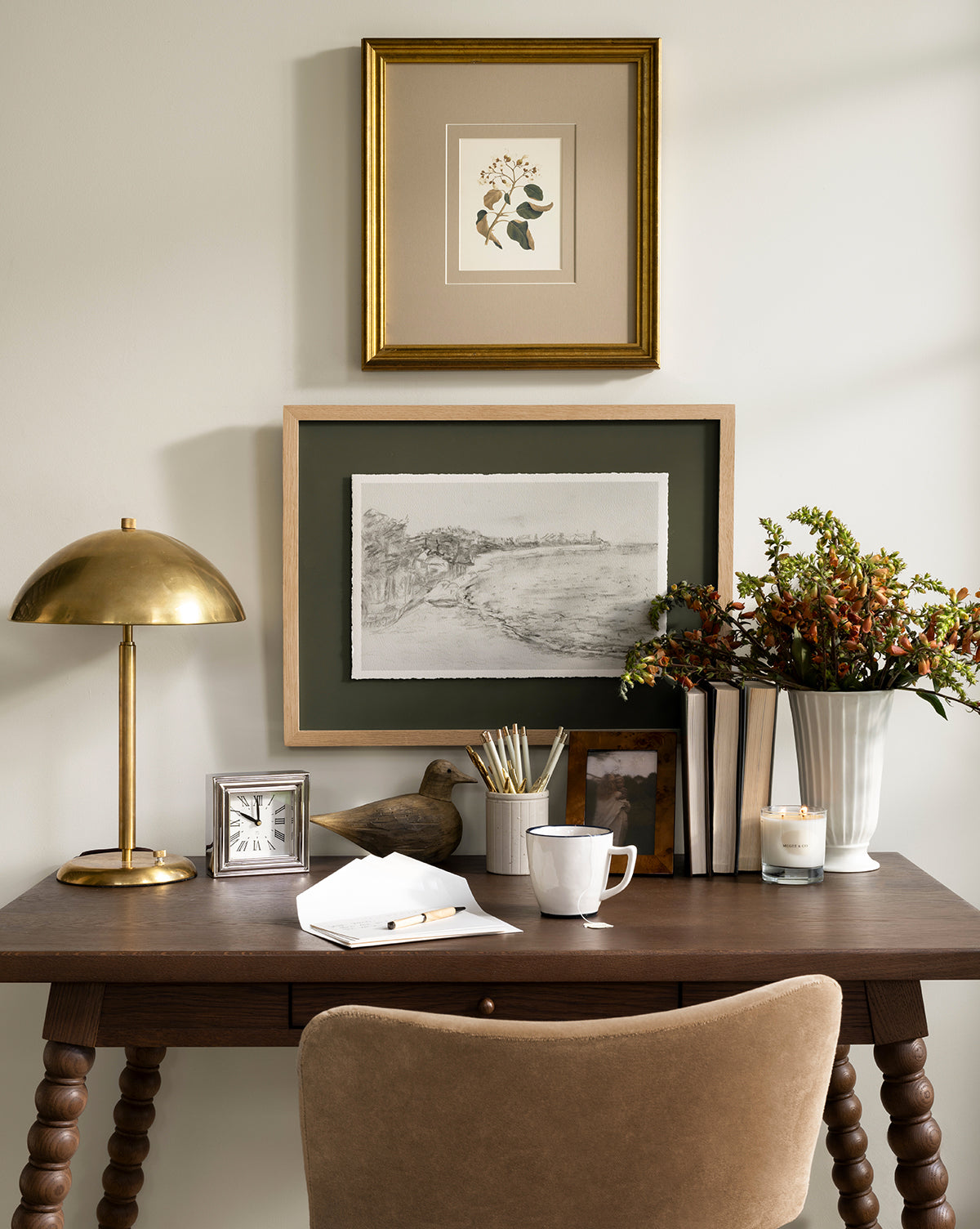 A tidy wooden desk with a beige chair, gold lamp, books, mug, and white vase with flowers adds charm to this decor. Lit candles glow while two Cove in Charcoal sketches in light oak frames hang above the desk, by McGee & Co.