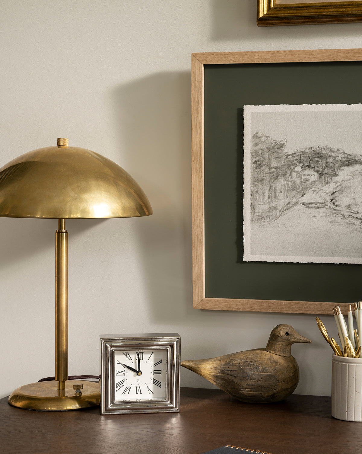 The Aldorno Desk Table Lamp, a silver clock, a wooden bird figurine, and a cup of pens rest on the desk, with a framed black-and-white sketch hanging above, by McGee & Co.