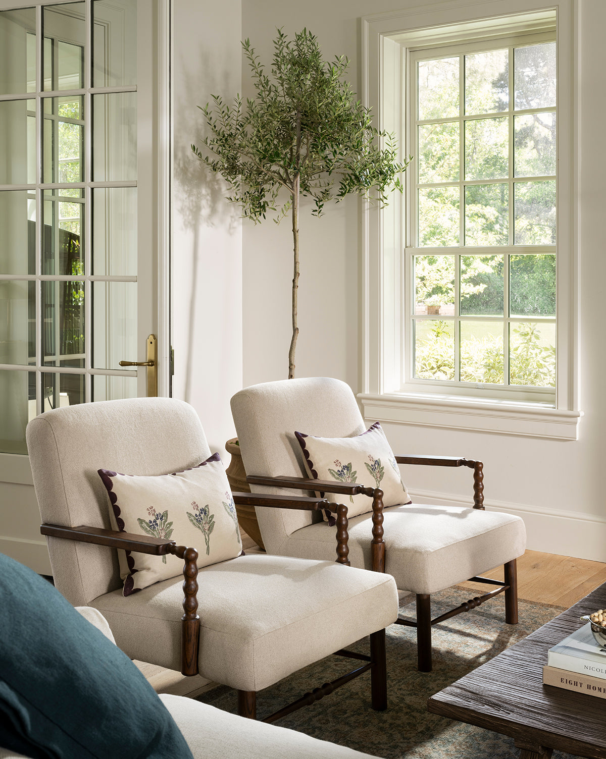 Two cream-colored Earnshaw Lounge Chairs with neutral upholstery and floral cushions sit by a large window beside a potted indoor tree. Sunlight fills the cozy living room, which also features a wooden coffee table in the foreground, by McGee & Co.