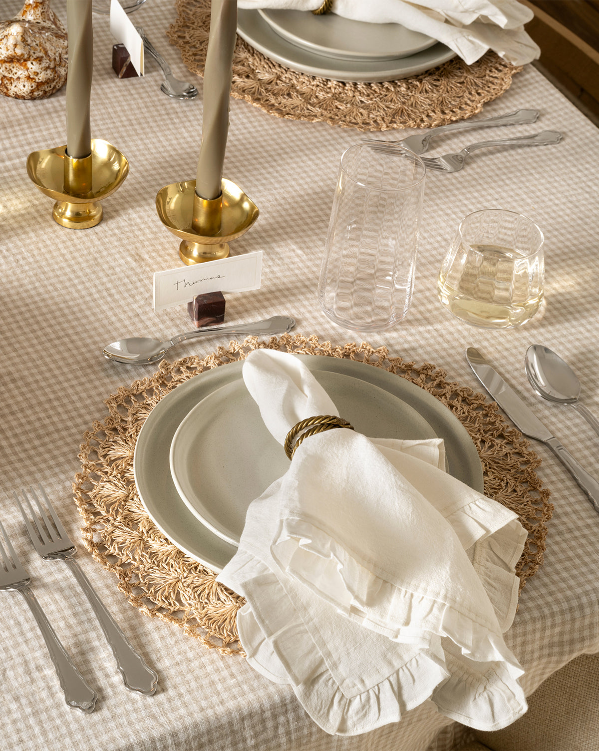 A neatly set dining table with a beige checkered tablecloth, woven placemats, stacked plates, Catania Linen Napkins (Set of 4) with gold rings, silver cutlery, clear glasses, brass candlesticks, and a name card by McGee & Co.