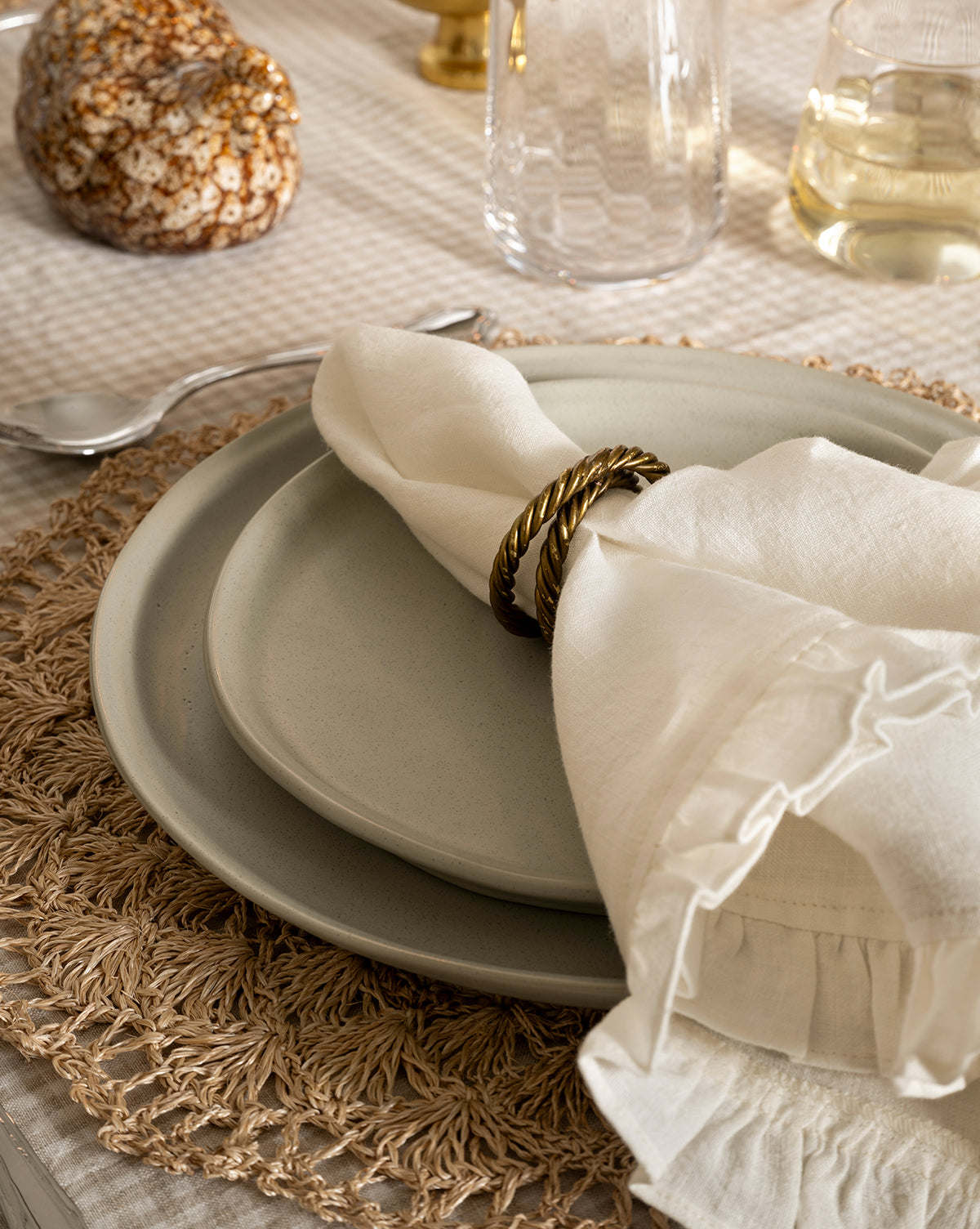 A close-up of a table setting featuring a cream-colored plate, the Aiden Soft Gray Side Plate, a linen napkin with a gold ring, and a woven placemat on a checkered tablecloth. A spoon, glasses, and decor are also visible by McGee & Co.